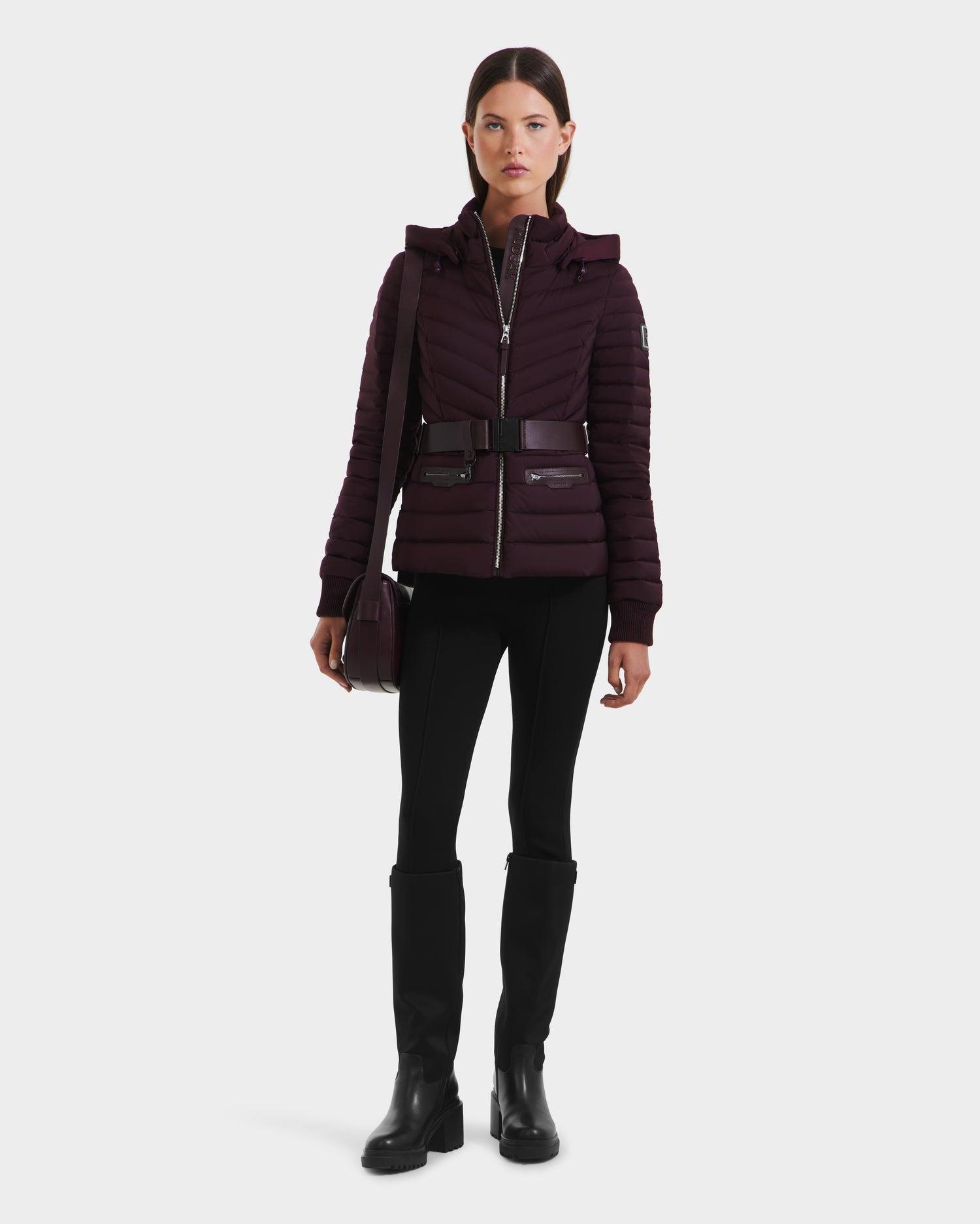 A woman with straight dark hair and a neutral expression faces forward, wearing the Rudsak SCARLETT M - 8125565 Bordeaux women’s lightweight down jacket with a vegan leather belt, styled with black pants, knee-high boots, and a matching crossbody bag.