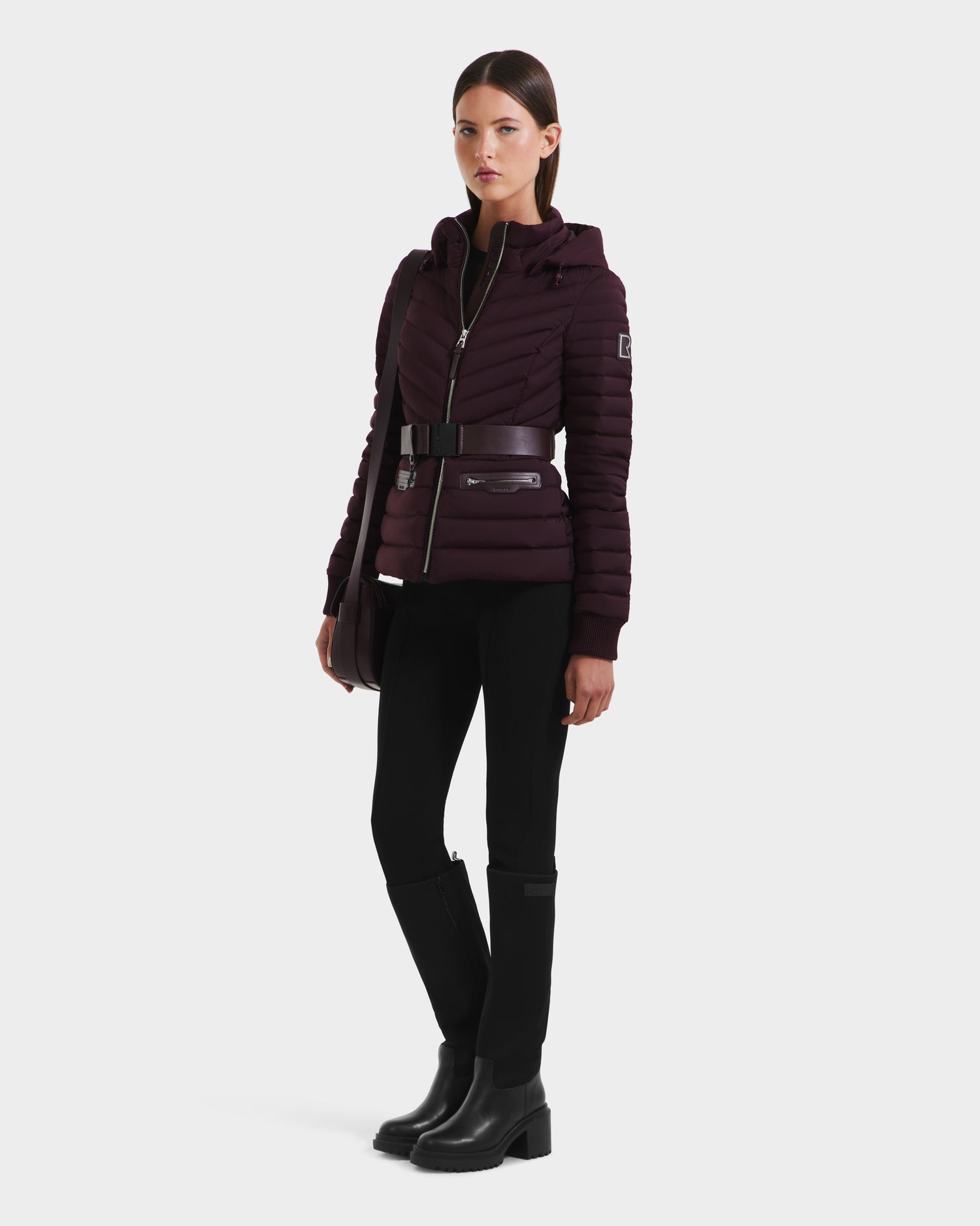 A woman poses against a white background wearing the Rudsak SCARLETT M - 8125565 Bordeaux fitted lightweight down jacket with a vegan leather belt, paired with black pants, knee-high boots, and a crossbody bag.