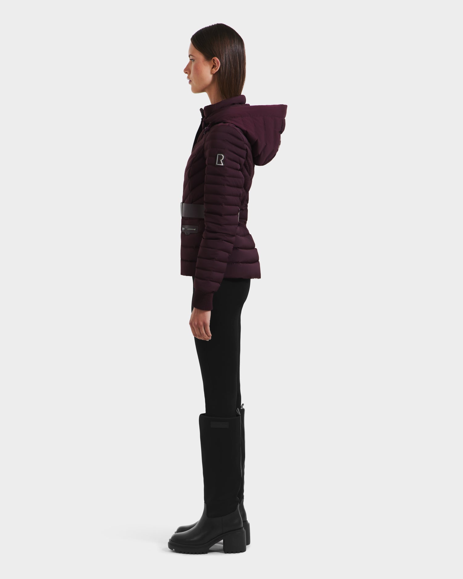 A woman in profile wears the Rudsak SCARLETT M - 8125565 Bordeaux puffer jacket with a water-resistant shell, black leggings, and knee-high black boots against a plain white background.