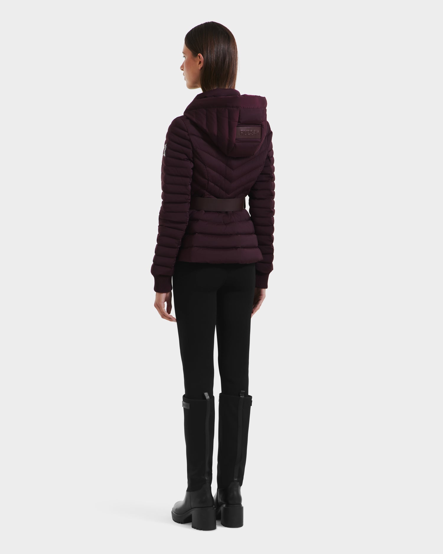 A woman faces away, wearing the Rudsak SCARLETT M - 8125565 Bordeaux, a dark purple women’s lightweight down jacket with a hood, paired with black pants and knee-high black boots against a plain white background.