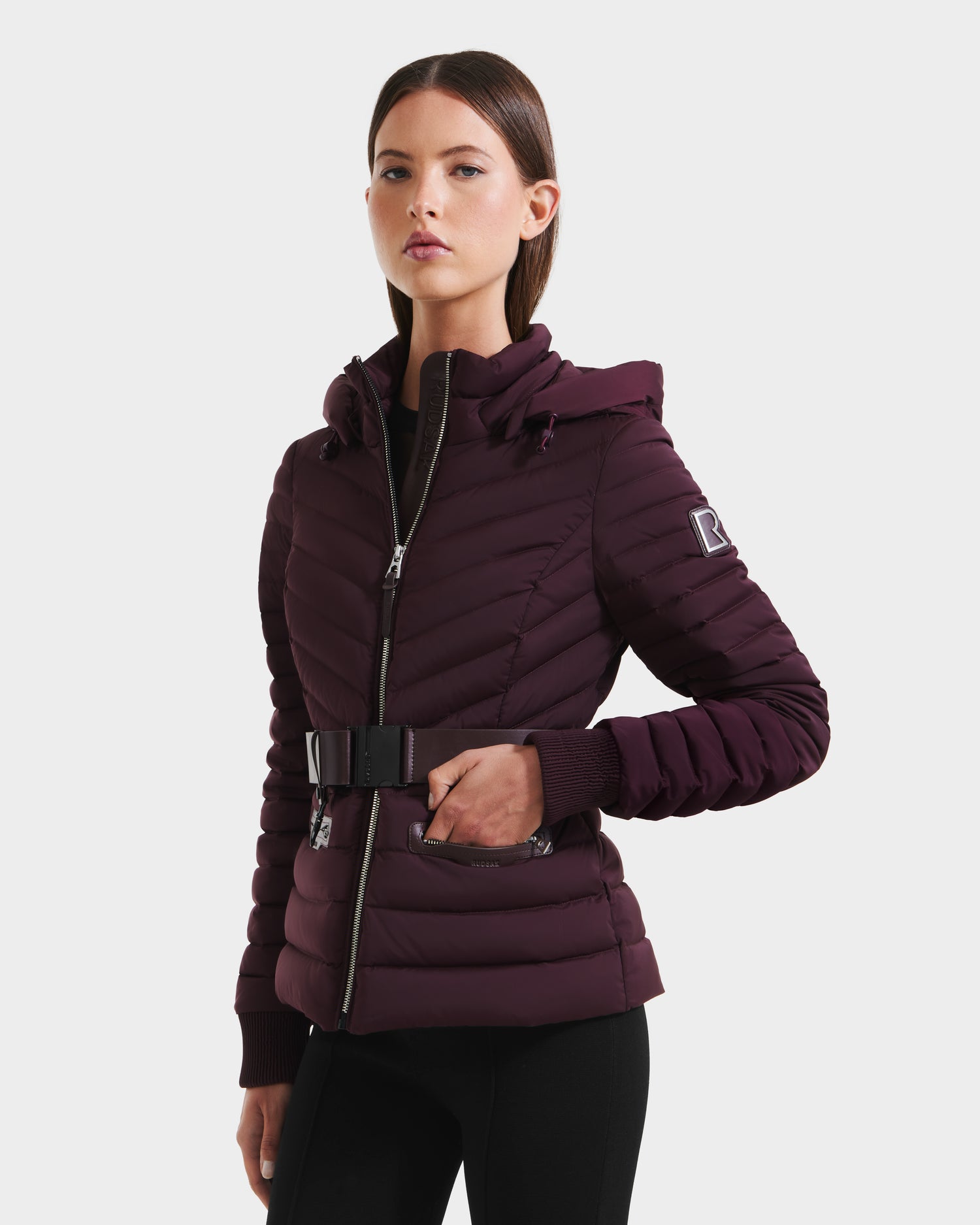 A woman models the Rudsak SCARLETT M - 8125565 Bordeaux puffer jacket, in dark purple with a hood and vegan leather waist belt, zipped up and styled with black pants, standing slightly angled against a plain white background.