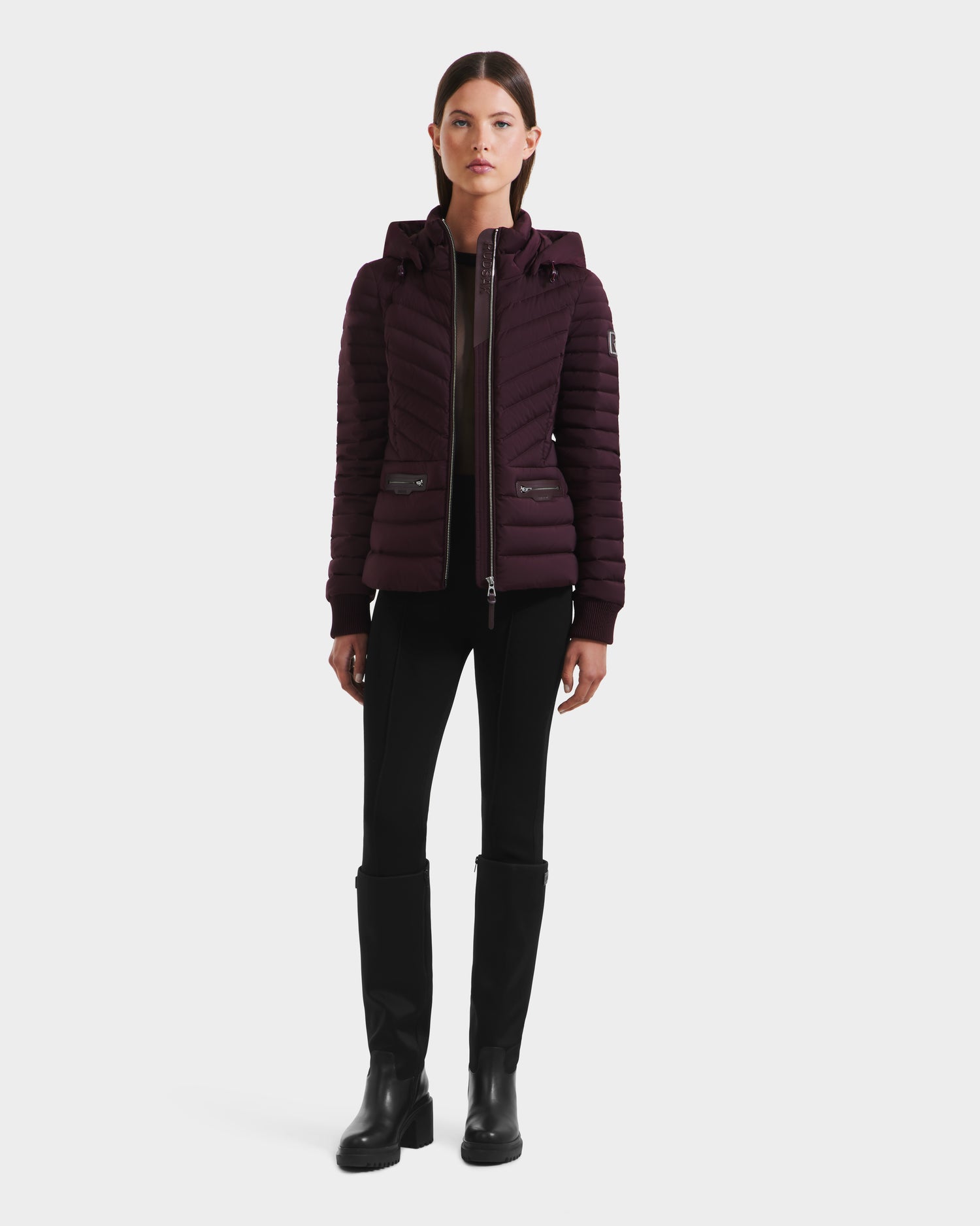 A woman stands facing forward wearing the Rudsak SCARLETT M - 8125565 Bordeaux lightweight down jacket, paired with black pants and knee-high boots, against a plain white background.