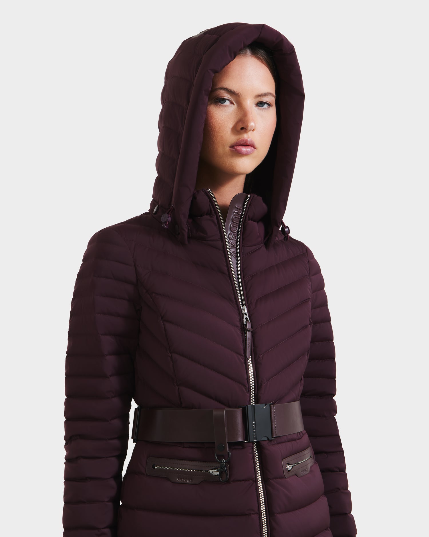 A woman wears the Rudsak SCARLETT M - 8125565 Bordeaux, a hooded, fitted dark burgundy lightweight down jacket with a zip closure and vegan leather belt, standing against a plain white background.