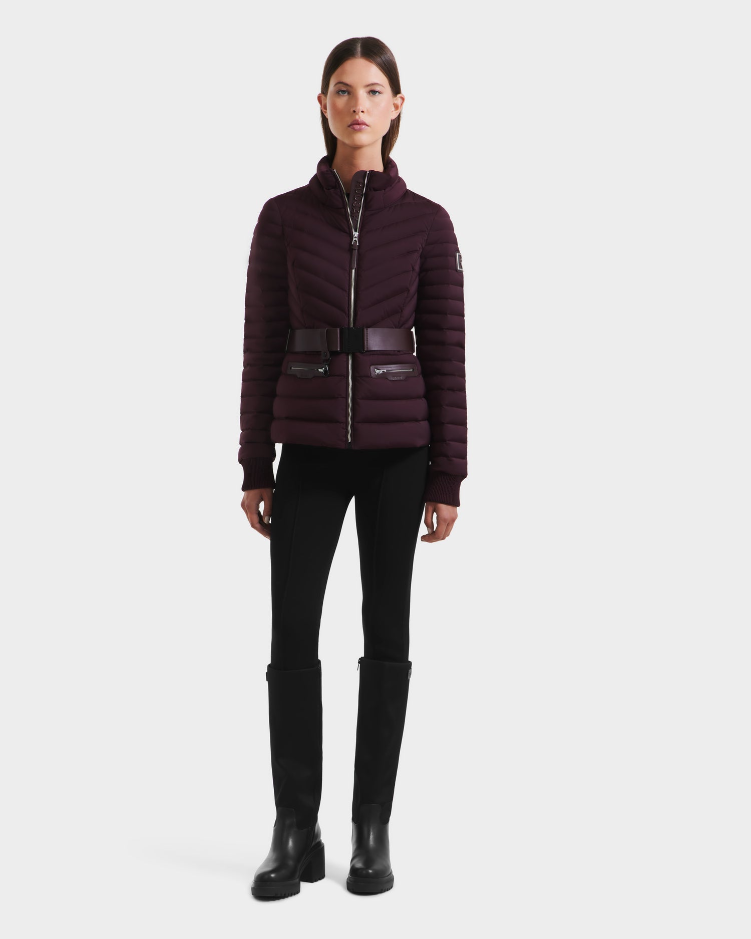 A woman stands facing forward wearing the Rudsak SCARLETT M - 8125565 Bordeaux fitted lightweight down jacket with a high collar, styled with black pants, a wide vegan leather belt, and knee-high black boots against a white background.