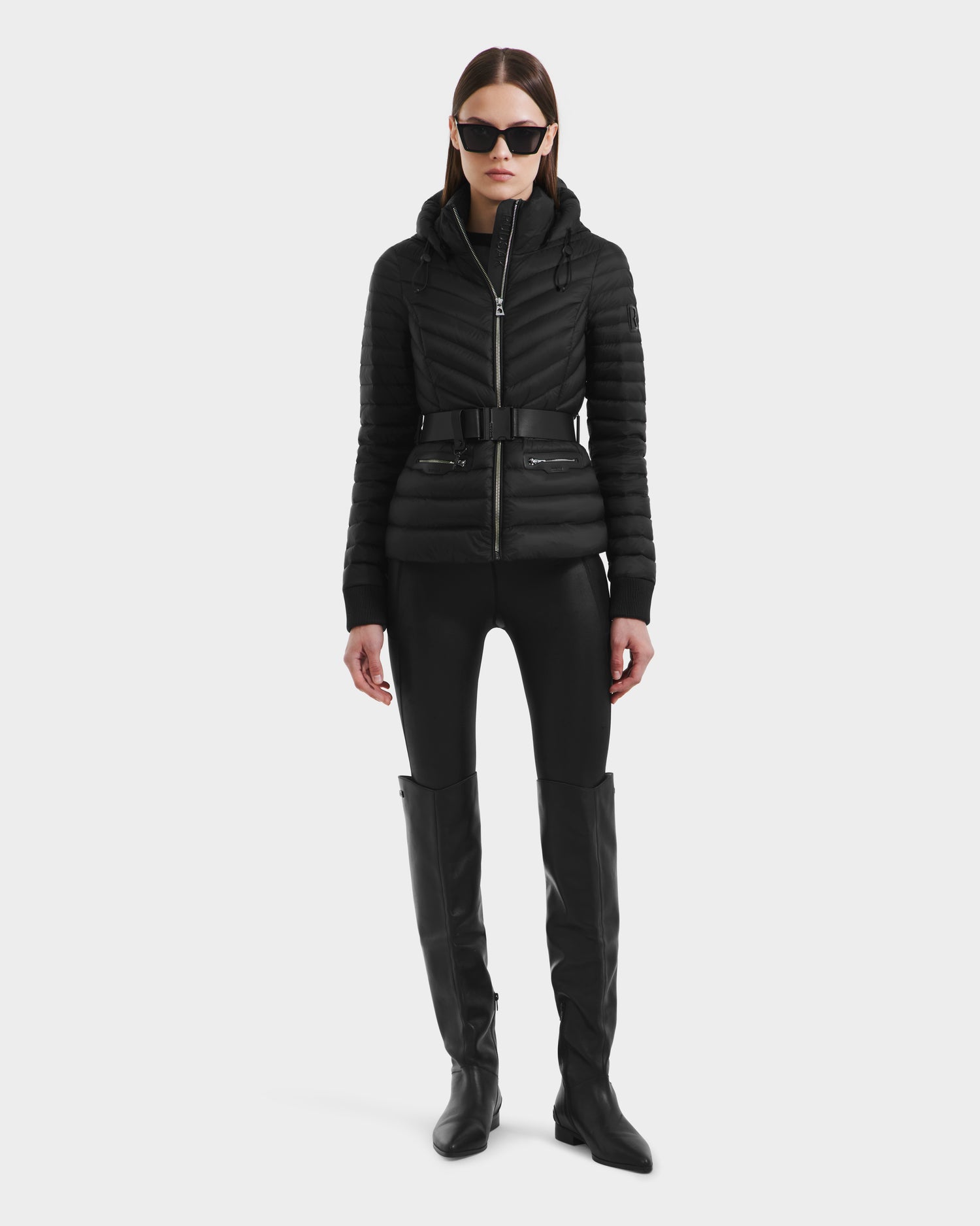 A woman wearing the Rudsak SCARLETT - 8125004 Black jacket, cinched with a vegan leather belt, along with black sunglasses, shiny black leggings, and knee-high boots stands against a plain white background.