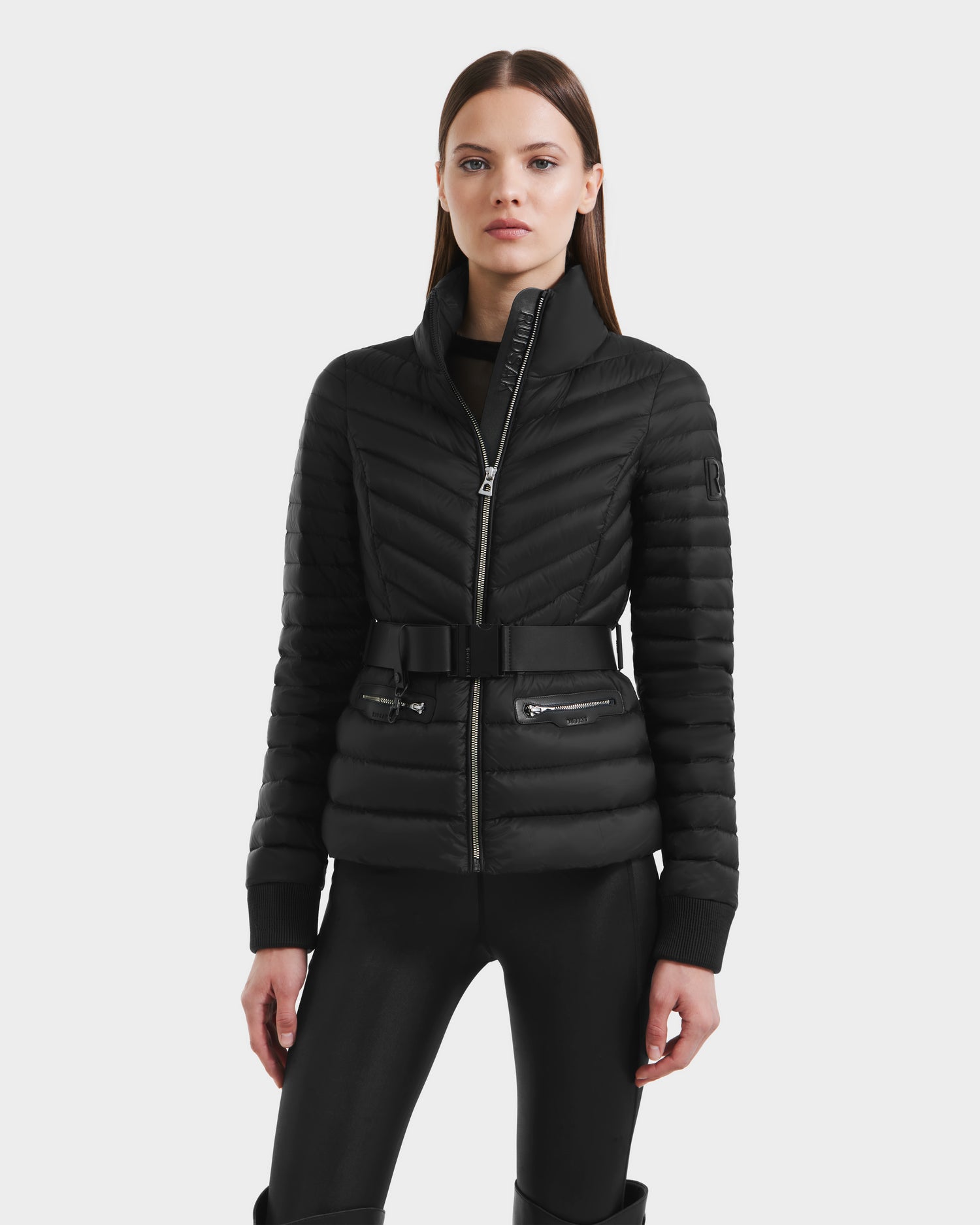 A woman stands facing forward against a plain white background, wearing the Rudsak SCARLETT - 8125004 Black fitted women’s lightweight down jacket with a front zipper, vegan leather belt, and zippered side pockets.