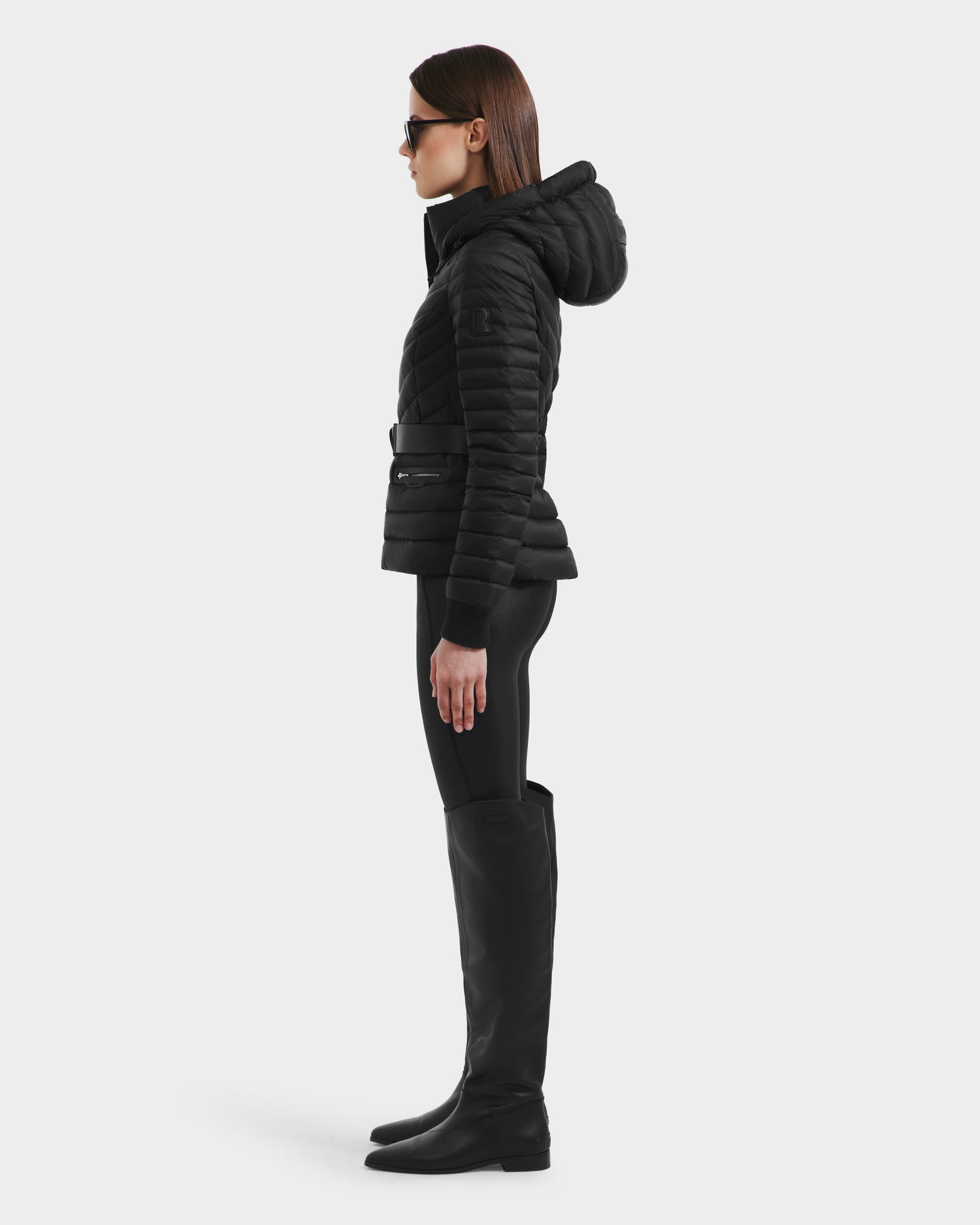 A woman wears the Rudsak SCARLETT - 8125004 Black coat, featuring a vegan leather belt and timeless elegance.