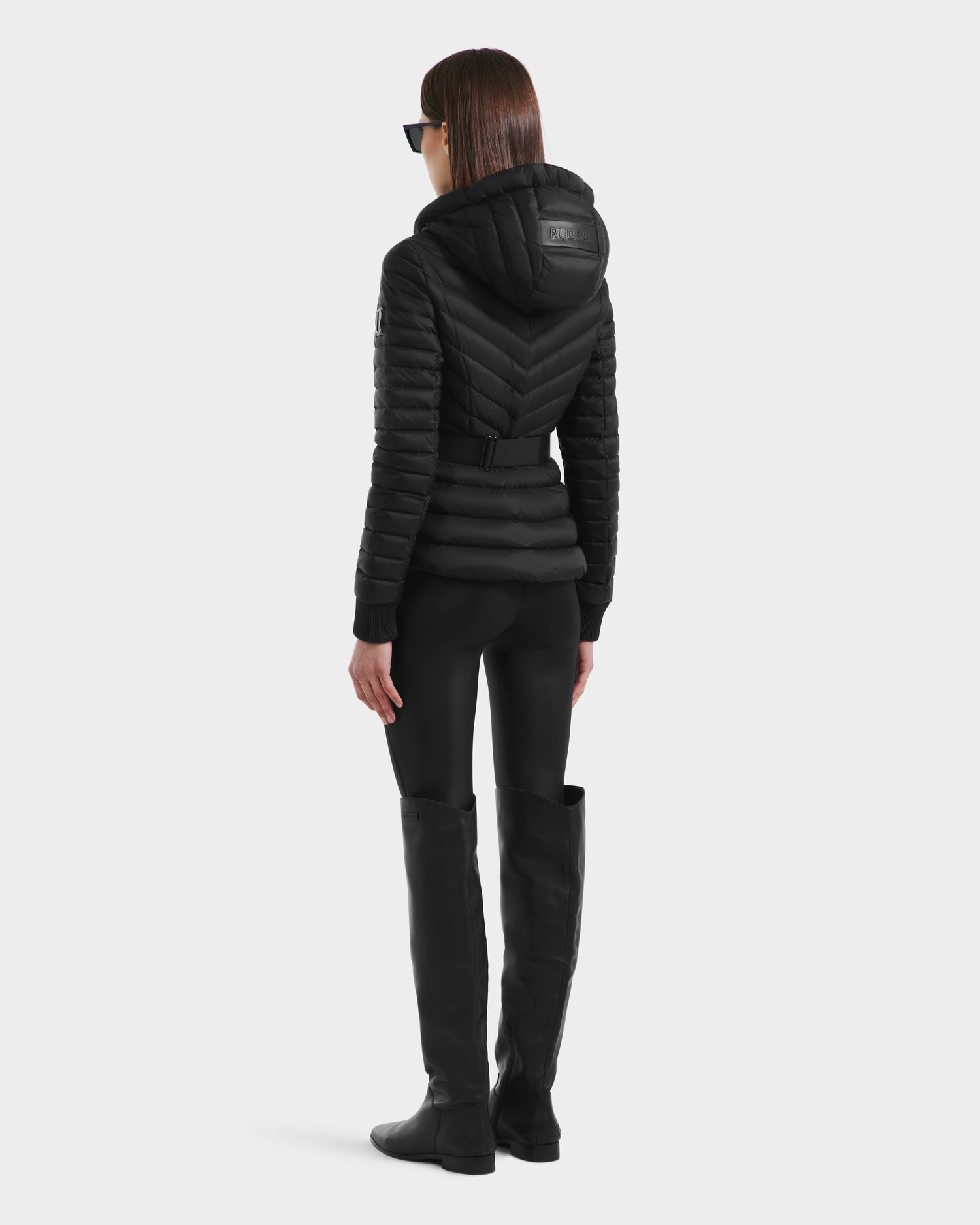 A person with straight brown hair, sunglasses, and black pants stands facing away in a Rudsak SCARLETT - 8125004 Black women’s lightweight down jacket with vegan leather belt and knee-high black boots against a plain light background.