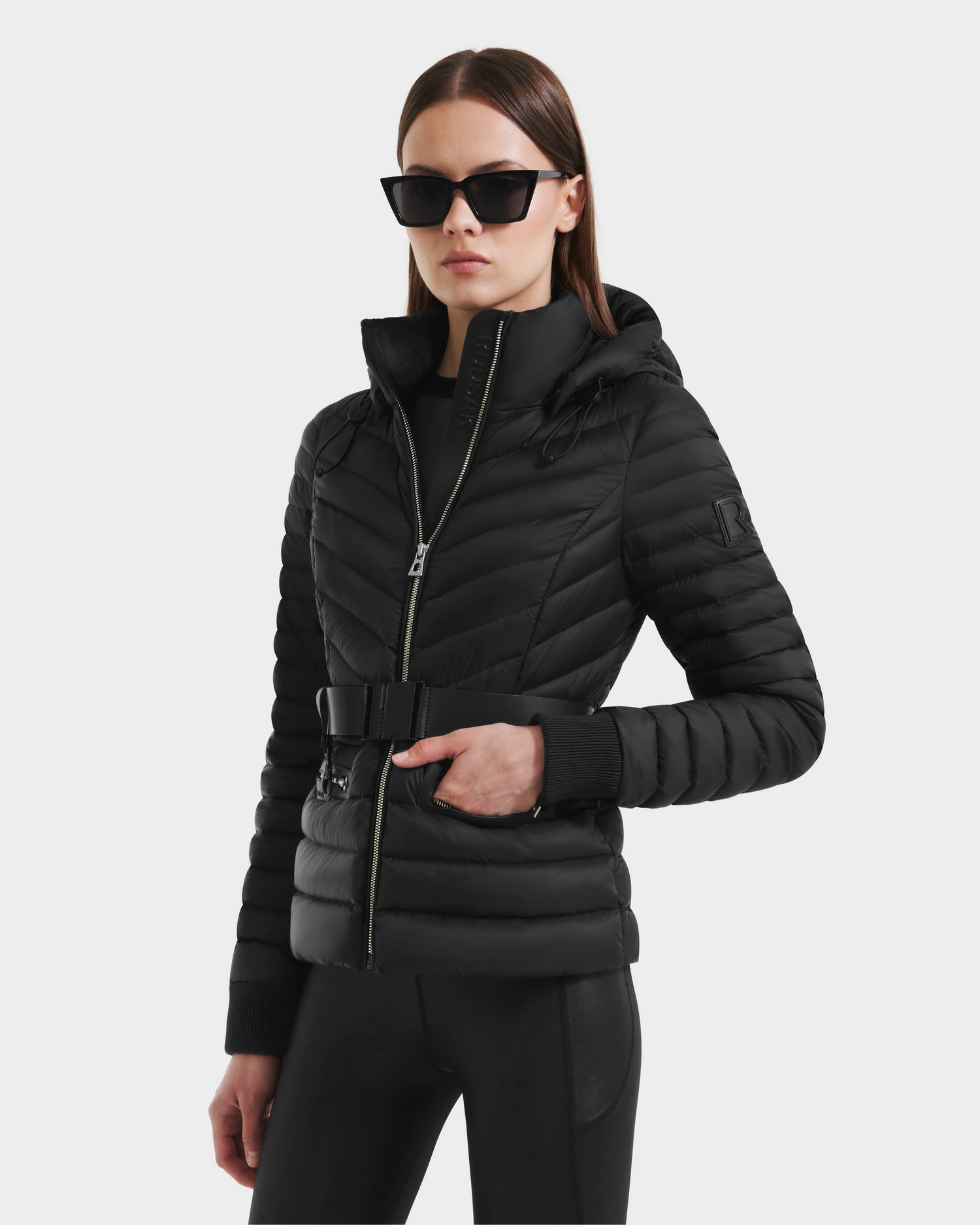 A woman in the Rudsak SCARLETT - 8125004 Black women’s lightweight down jacket, black pants, and large black sunglasses poses against a plain light background.