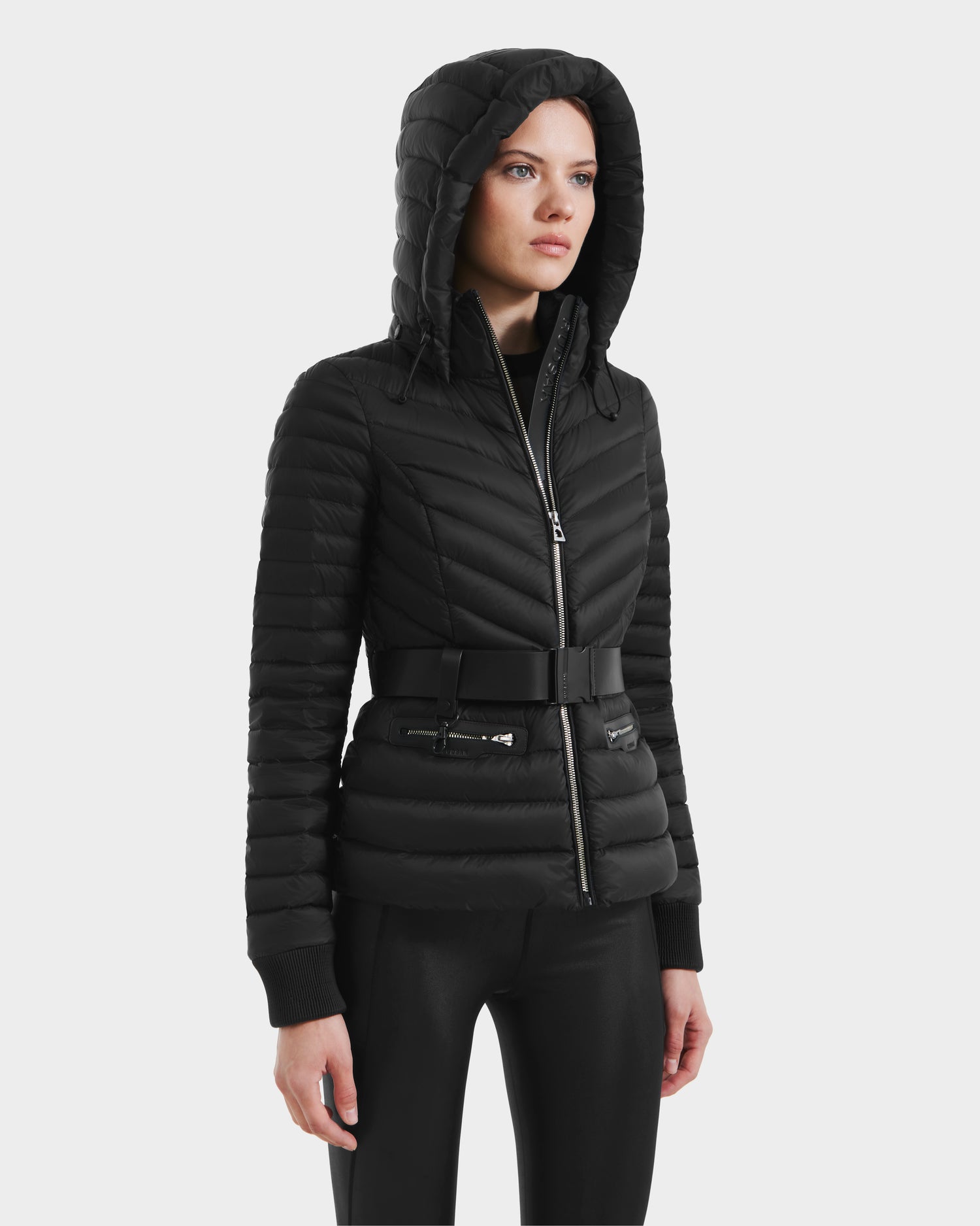A woman models the Rudsak SCARLETT - 8125004 Black jacket, featuring a black quilted design, hood, and vegan leather waist belt, standing against a plain white background with her hands relaxed at her sides.