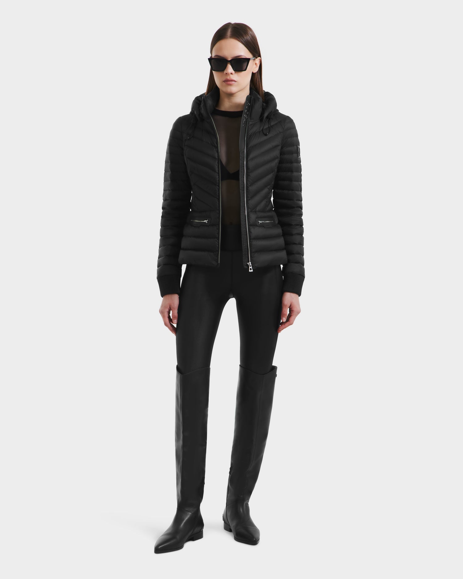 A woman wearing the Rudsak SCARLETT - 8125004 Black lightweight down jacket with a vegan leather belt, paired with black mesh top, pants, knee-high boots, and dark sunglasses stands against a plain white background.