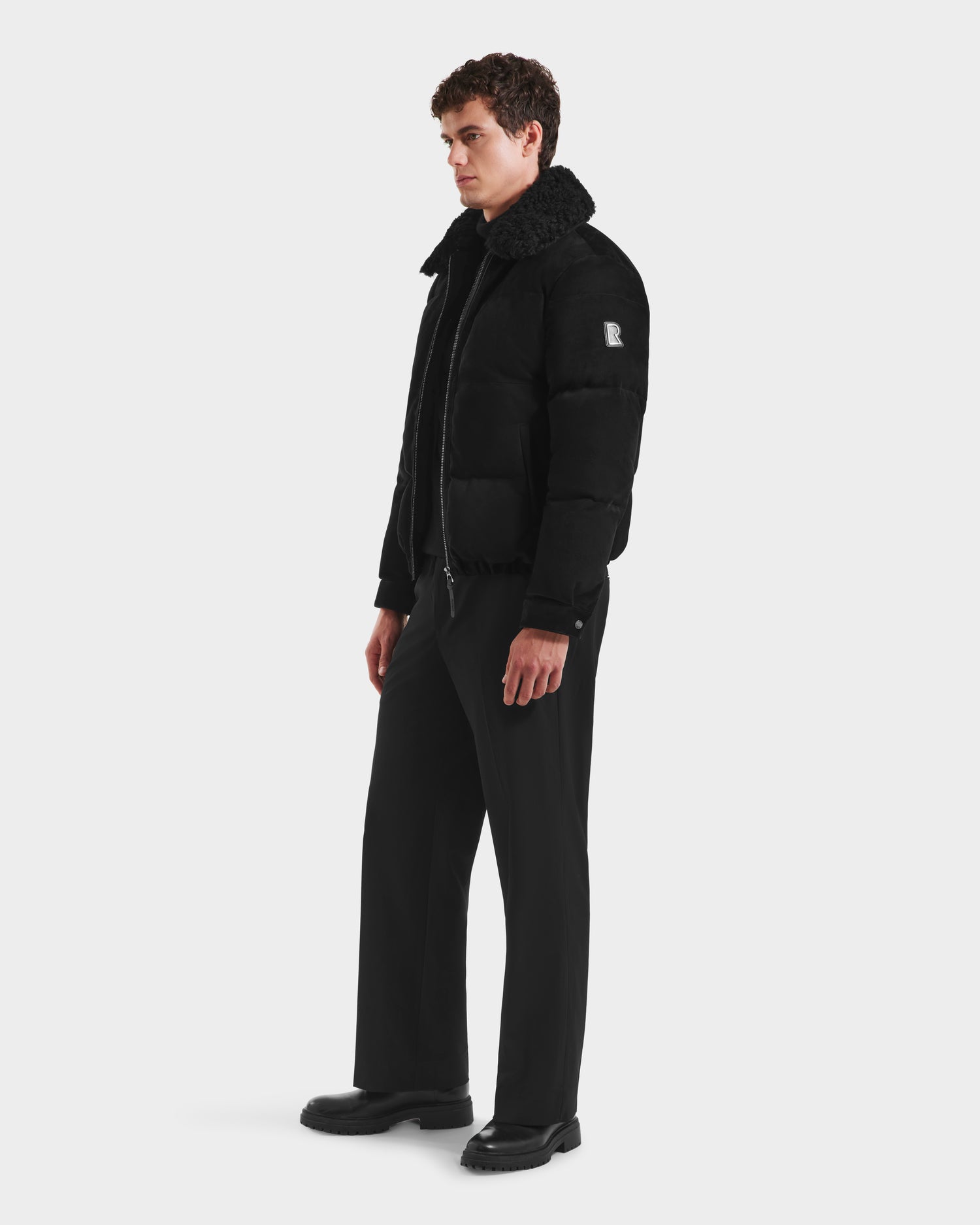 A man stands at an angle in front of a white background, wearing the Rudsak SCOTT - 6125685 Black shearling-collar puffer jacket with black pants and shoes.