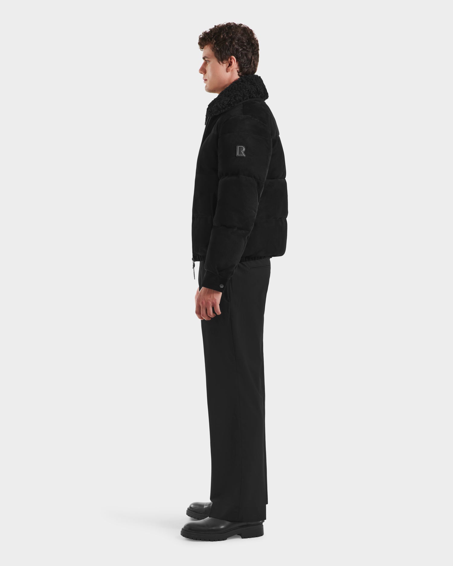 A man in profile wears the Rudsak SCOTT - 6125685 Black aviator bomber jacket with a shearling collar, black pants, and black shoes against a plain white background. The jacket features a patch on the left upper arm.