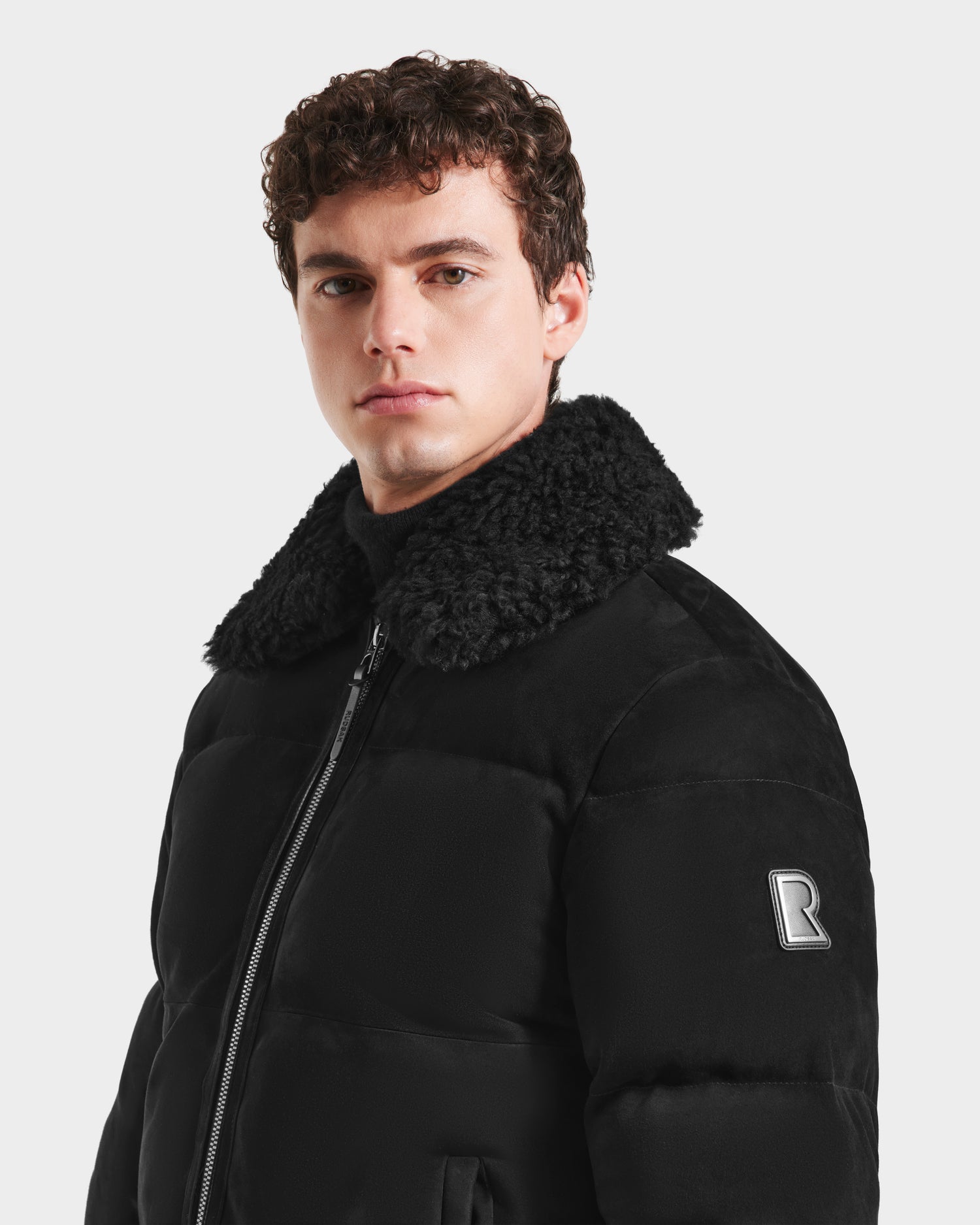 A young man with curly brown hair wears the Rudsak SCOTT - 6125685 Black men’s aviator bomber jacket with a shearling collar and bold R logo on the sleeve, standing against a plain light background.