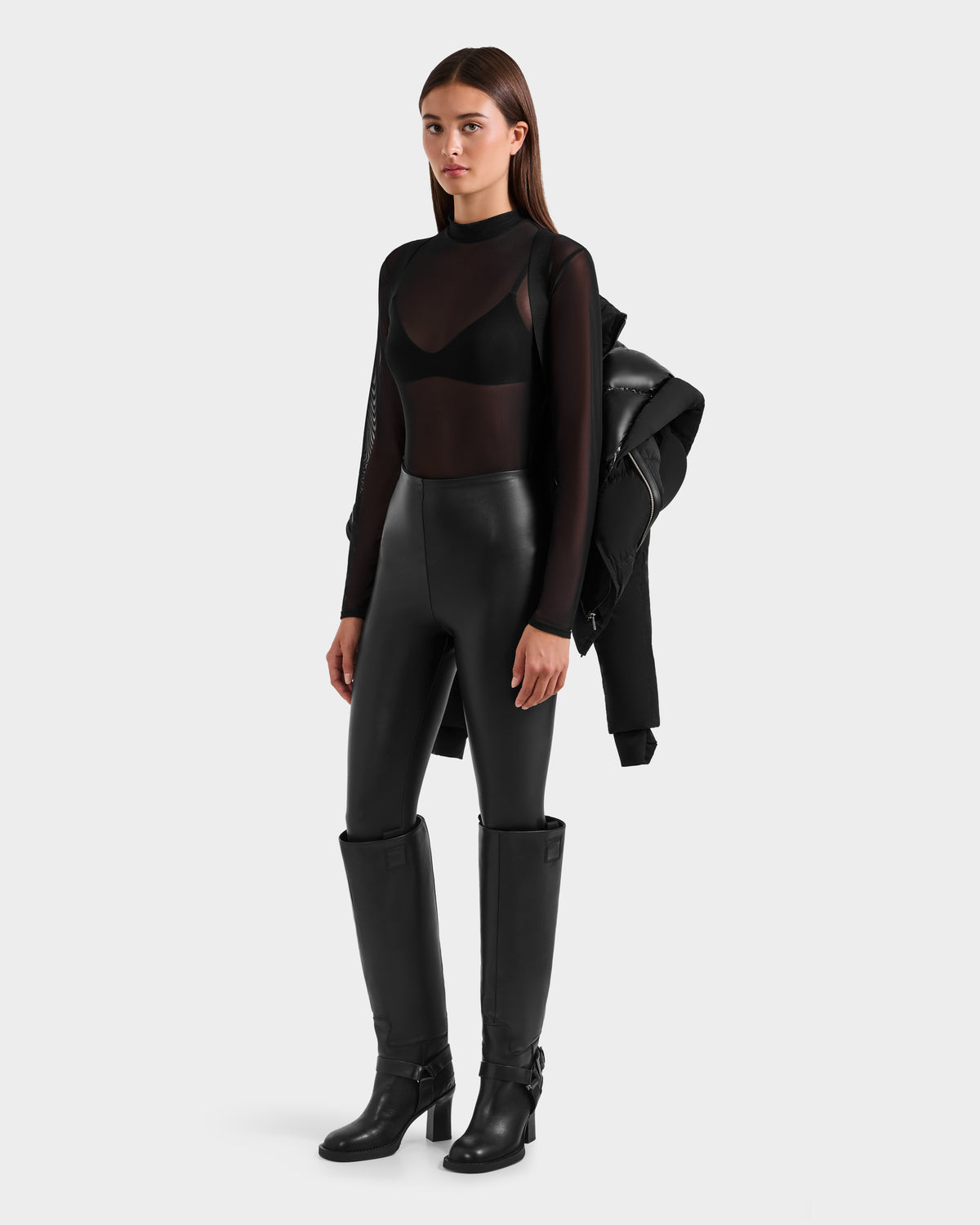 A woman poses against a plain background in high-waisted black leather pants, heeled knee-high boots, and a sheer black long-sleeve top over a bra, holding the Rudsak SHALI - 8124588 Black winter jacket over one shoulder.