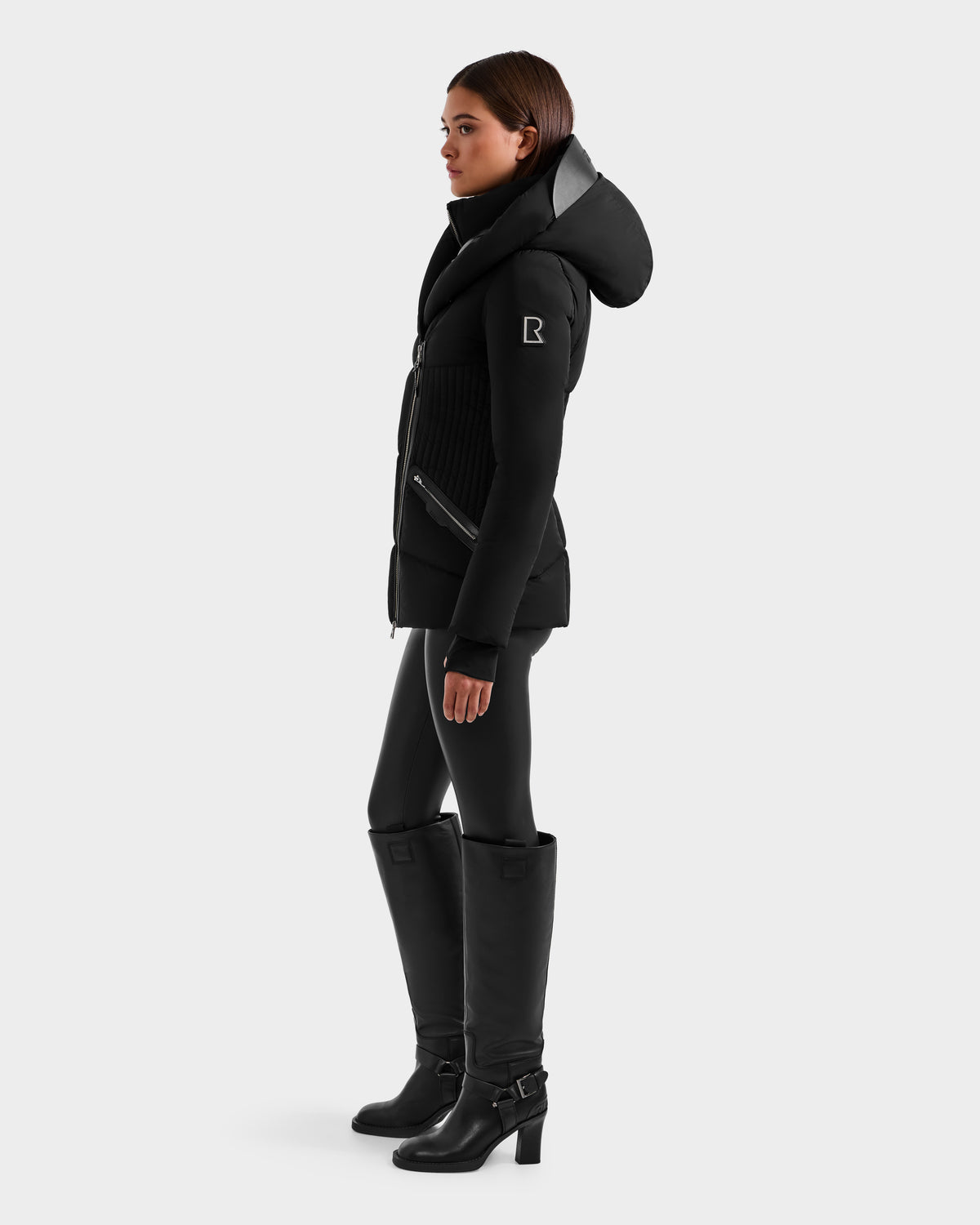 A woman in profile wears the Rudsak SHALI - 8124588 Black fitted winter jacket with hood, styled with black leggings and knee-high heeled, buckled boots, set against a plain light gray background.