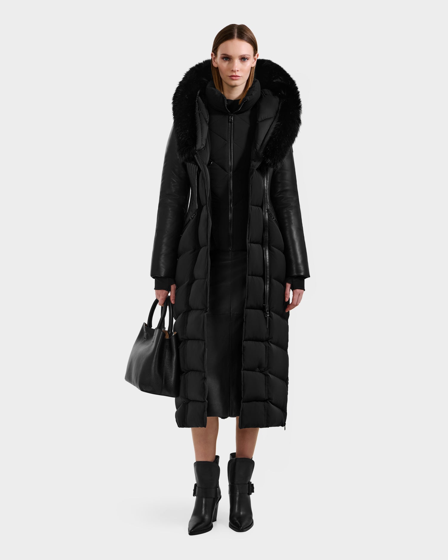 A woman faces forward in the Rudsak SHANNON - 8121511 BLACK quilted maxi down parka with leather details, styled with a black dress, ankle boots, and a black handbag against a plain white background.