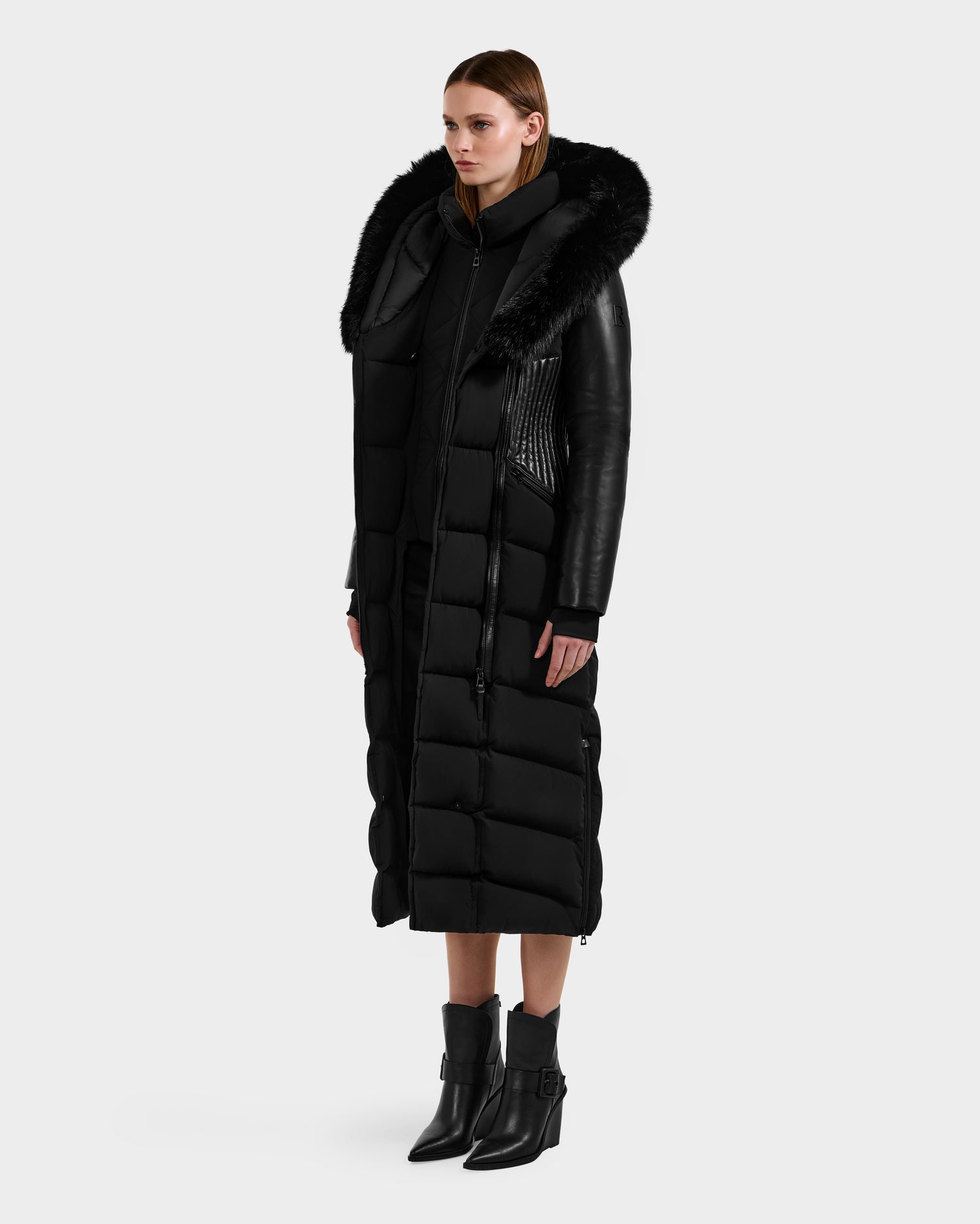 A woman stands in the SHANNON - 8121511 BLACK quilted maxi down parka by Rudsak, featuring leather details, a large fur-trimmed hood, and black ankle boots. The RDS-certified coat contrasts with the plain light gray background.