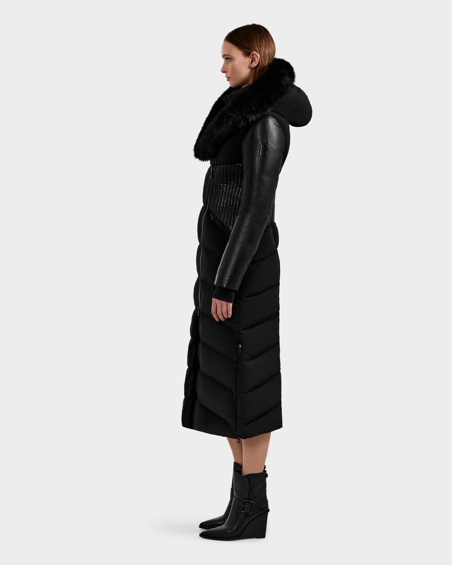 A woman stands in profile wearing the Rudsak SHANNON - 8121511 BLACK quilted maxi down parka with a fur collar, hood, and leather-detailed sleeves. She pairs it with heeled black ankle boots against a plain light background.