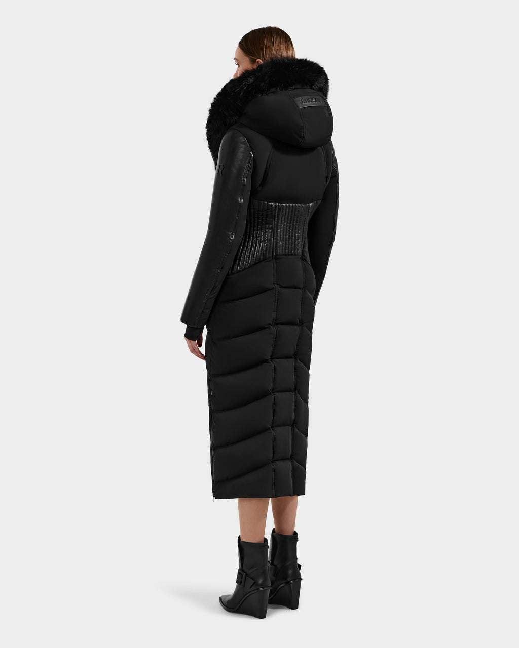 A woman with short brown hair faces away, wearing the Rudsak SHANNON - 8121511 BLACK puffer coat with a fur-trimmed hood and leather sleeves, paired with black heeled ankle boots that emphasize the coat’s tailored waist.