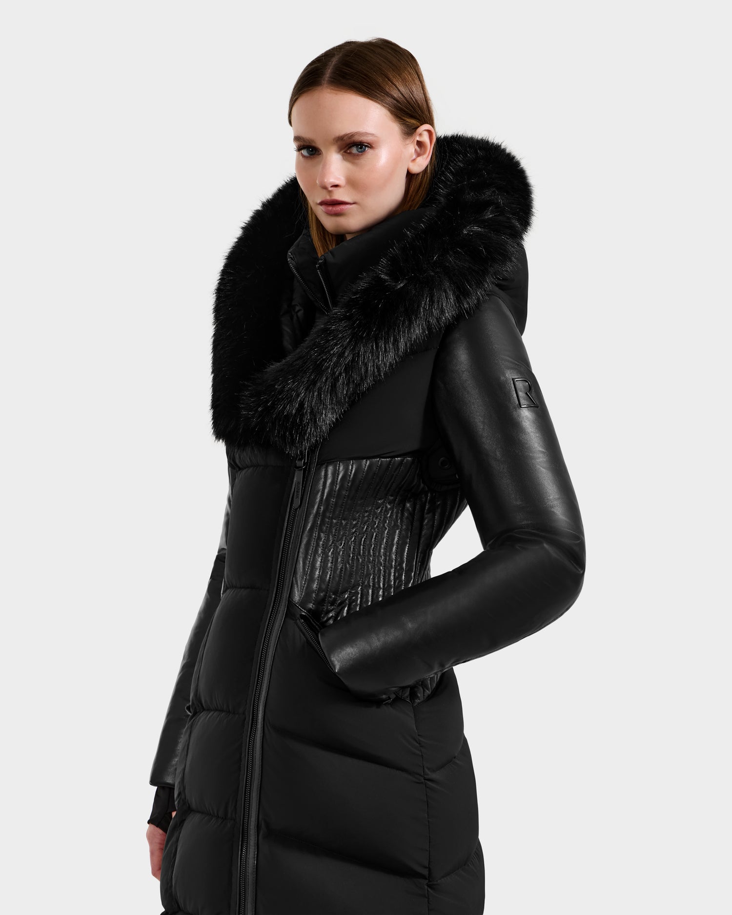 A woman stands confidently against a plain background wearing the Rudsak SHANNON - 8121511 BLACK, a stylish black quilted maxi down parka with leather details and a plush faux fur collar.