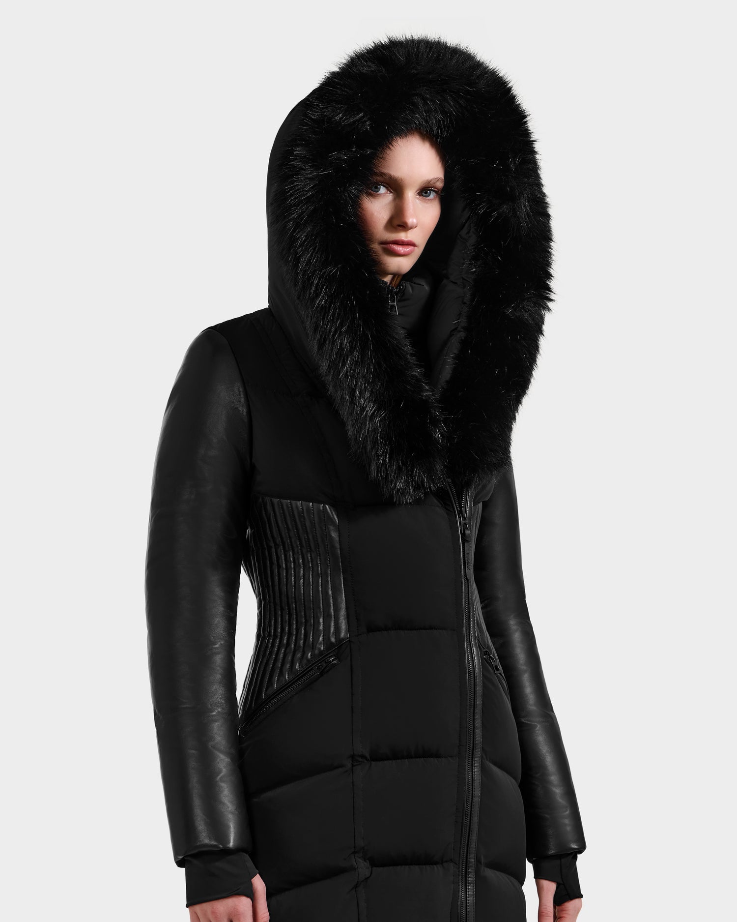 A person wears the Rudsak SHANNON - 8121511 BLACK, a long black hooded quilted maxi down parka with wide faux fur trim, leather-like sleeves, and quilted panels, standing against a plain light gray background.