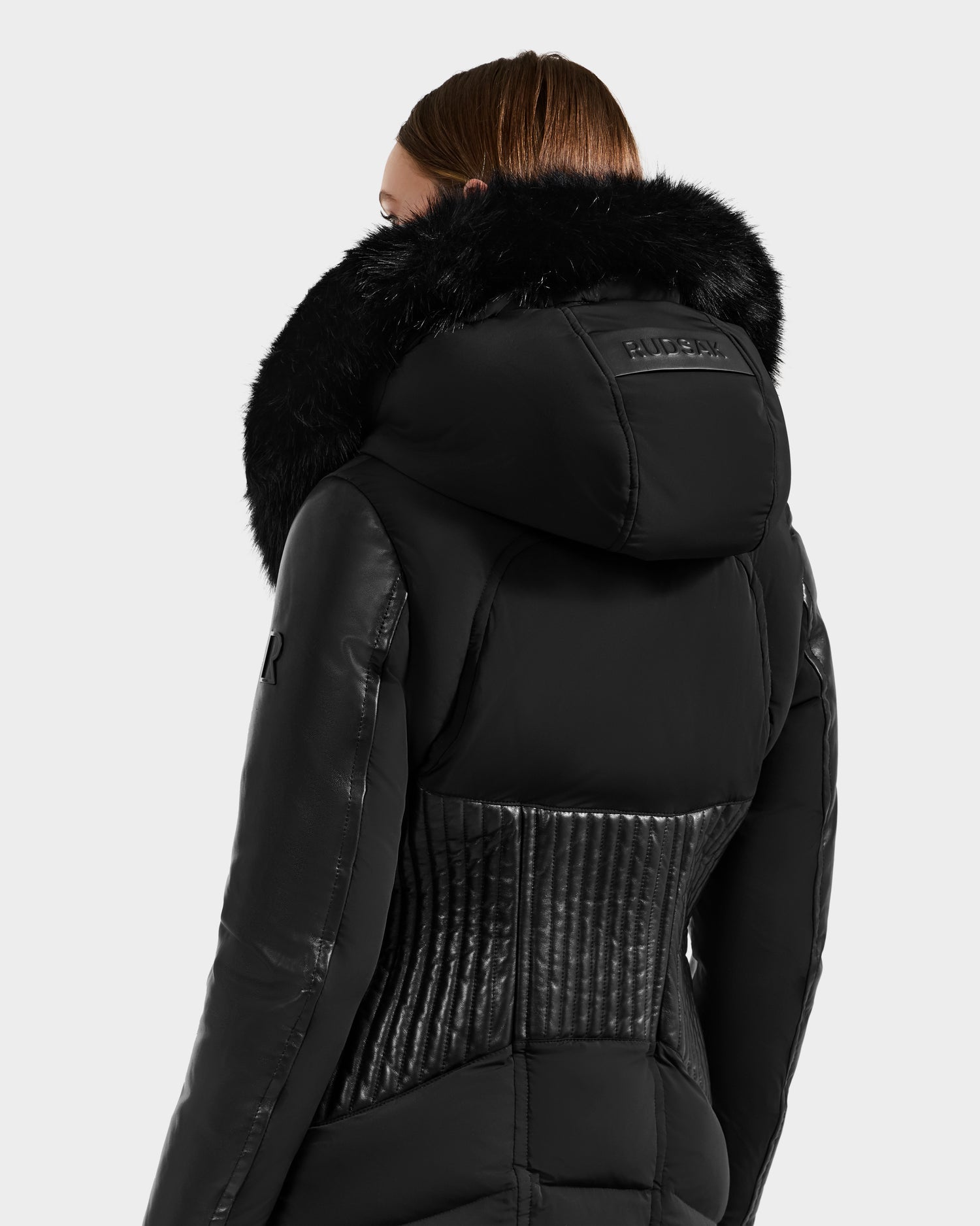 A person with straight brown hair models the Rudsak SHANNON - 8121511 BLACK fitted puffer jacket with quilted details and a large faux fur-trimmed hood, shown from the back against a plain light gray background.