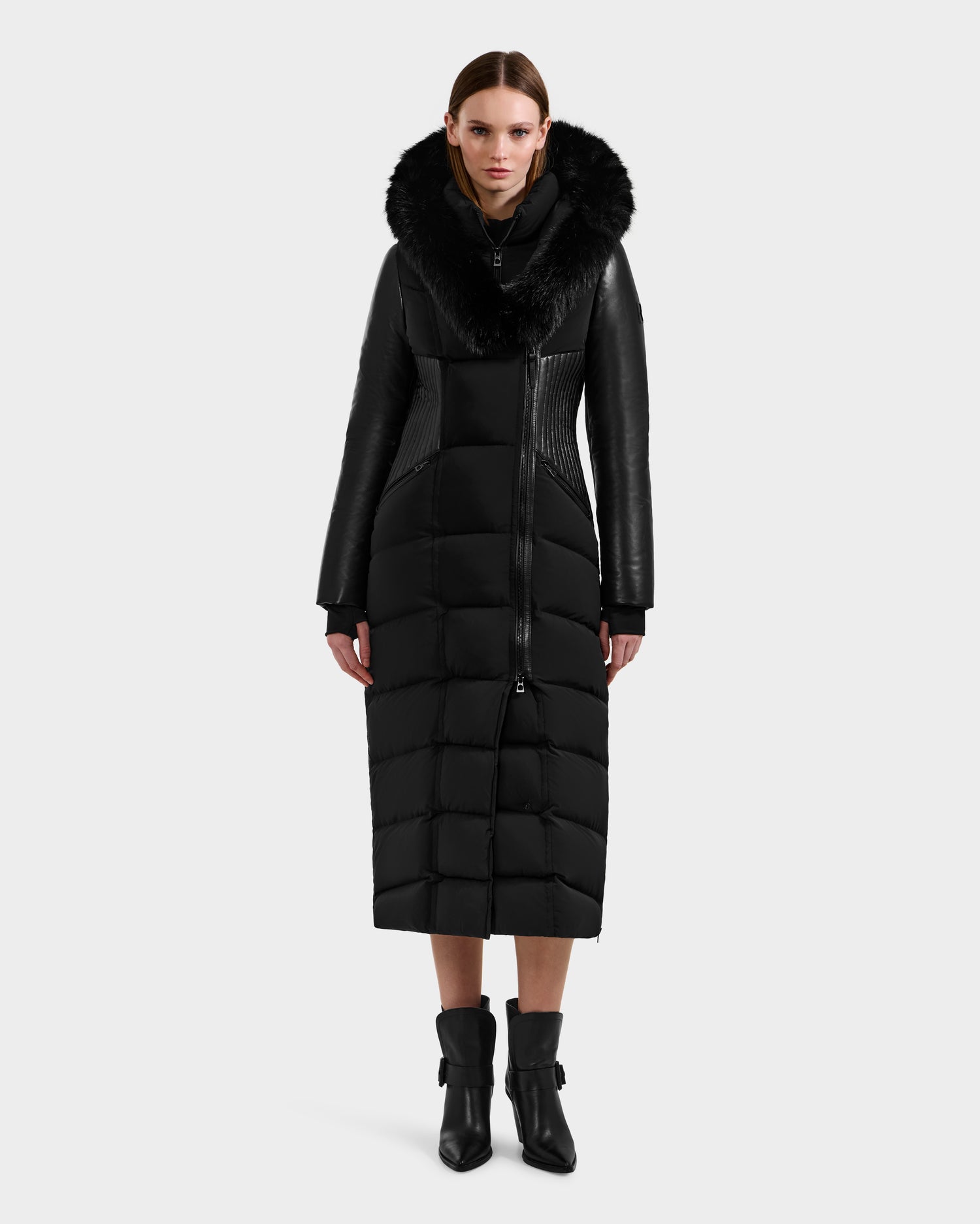A woman stands facing forward in the Rudsak SHANNON - 8121511 BLACK, a long black quilted maxi down parka with a fur-trimmed hood and leather-like sleeves. She pairs it with black ankle boots and wears her hair slicked back for a sleek look.
