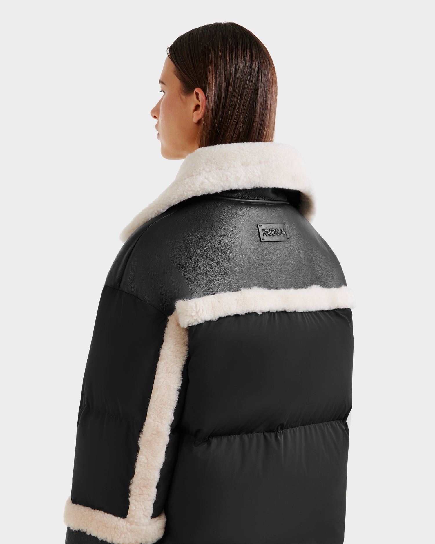 A woman with straight brown hair models the Rudsak SHIRLEY - 8125613 Black women’s down jacket with white shearling trim and collar, facing slightly away from the camera against a plain background.