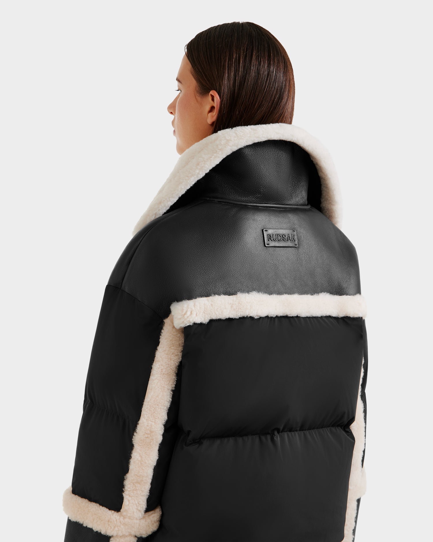 A woman with straight brown hair wears the Rudsak SHIRLEY - 8125613 Black shearling jacket with white sherpa trim and a large collar, revealing a visible RUDSAK patch on the back as she faces away from the camera.