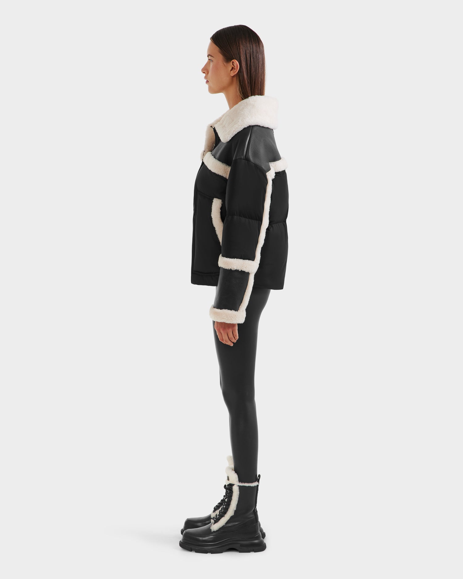 A woman stands in profile against a white background, wearing the Rudsak SHIRLEY - 8125613 Black women’s down jacket with white shearling trim—perfect winterwear.