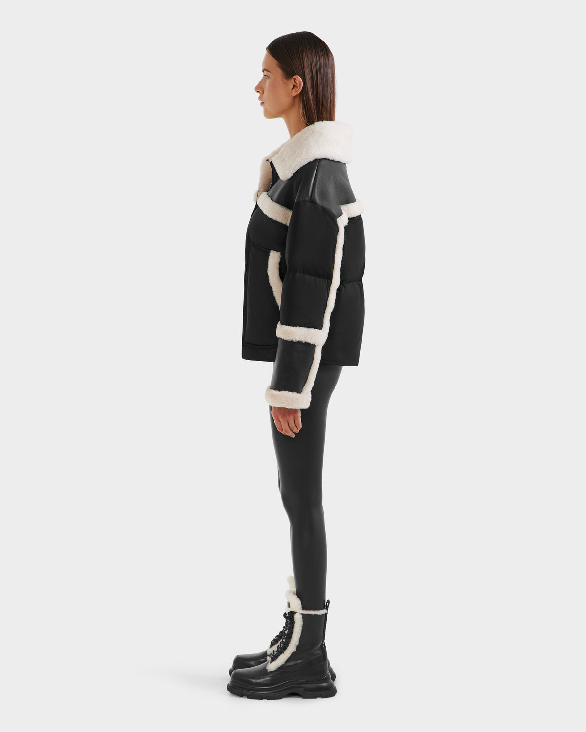 A person in profile showcases chic winter style wearing Rudsak’s SHIRLEY - 8125613 Black shearling jacket, glossy black leggings, and black lace-up boots with white trim, all set against a plain white background.
