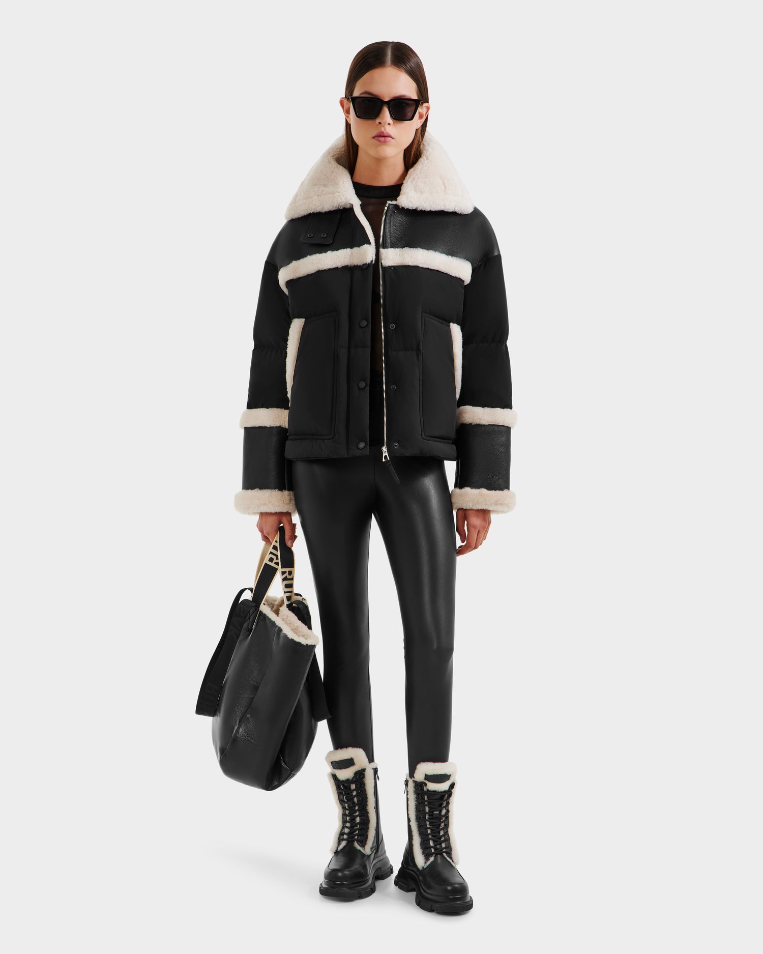 A woman wearing Rudsaks SHIRLEY - 8125613 Black shearling jacket, black sunglasses, faux leather pants, lace-up boots with white trim, and holding a large black bag stands against a plain white background.