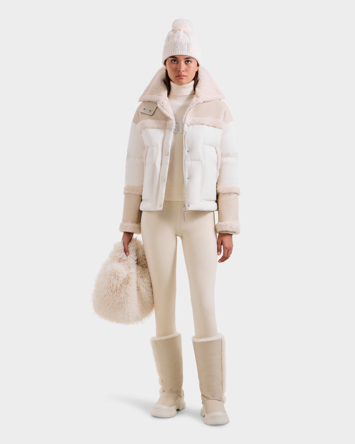 A woman stylishly poses against a light backdrop wearing Rudsak’s SHIRLEY - 8125613 Vanilla women’s down jacket, paired with fitted pants, fur-trimmed boots, a pom-pom knit hat, and holding a fluffy beige bag.