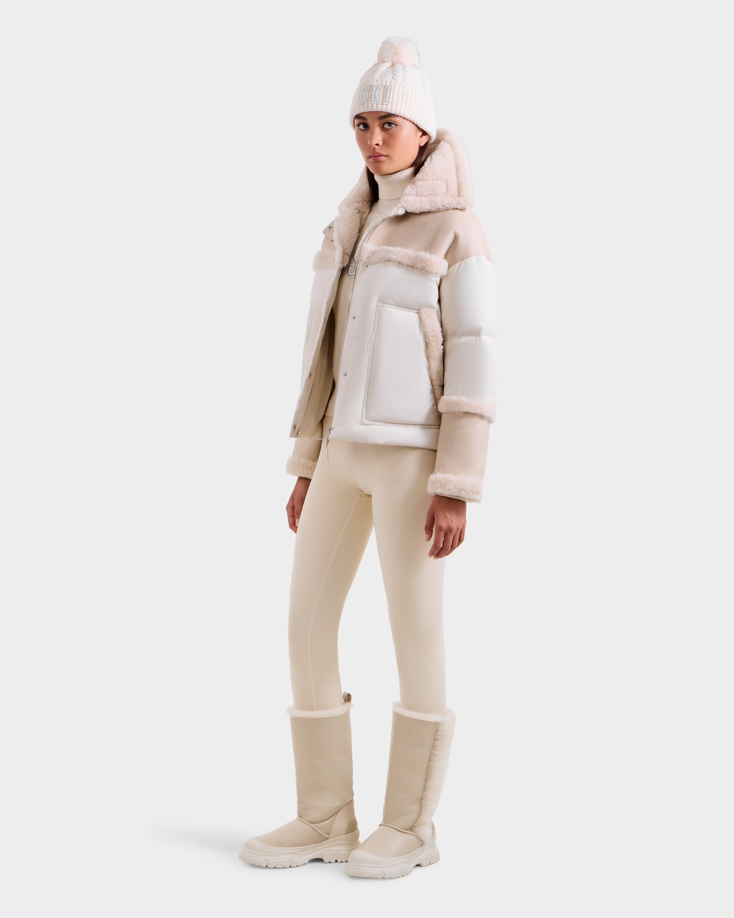 A woman models the Rudsak SHIRLEY - 8125613 Vanilla down jacket with faux fur trim, paired with a pom-pom beanie, fitted pants, and tall insulated boots, all in cream and white, against a plain white background.