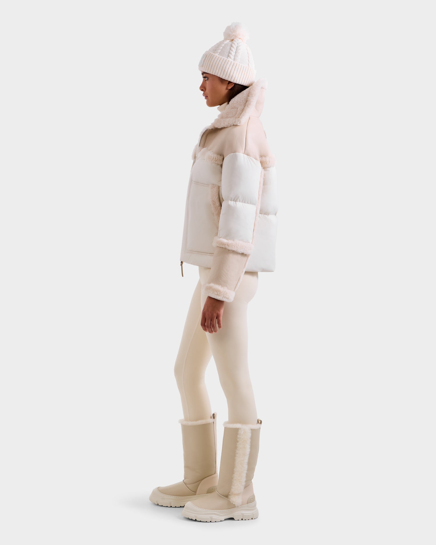 A person stands sideways in cozy winterwear, featuring the Rudsak SHIRLEY - 8125613 Vanilla women’s down jacket, a white knit pom-pom hat, cream leggings, and beige fur-trim knee-high boots against a plain background.