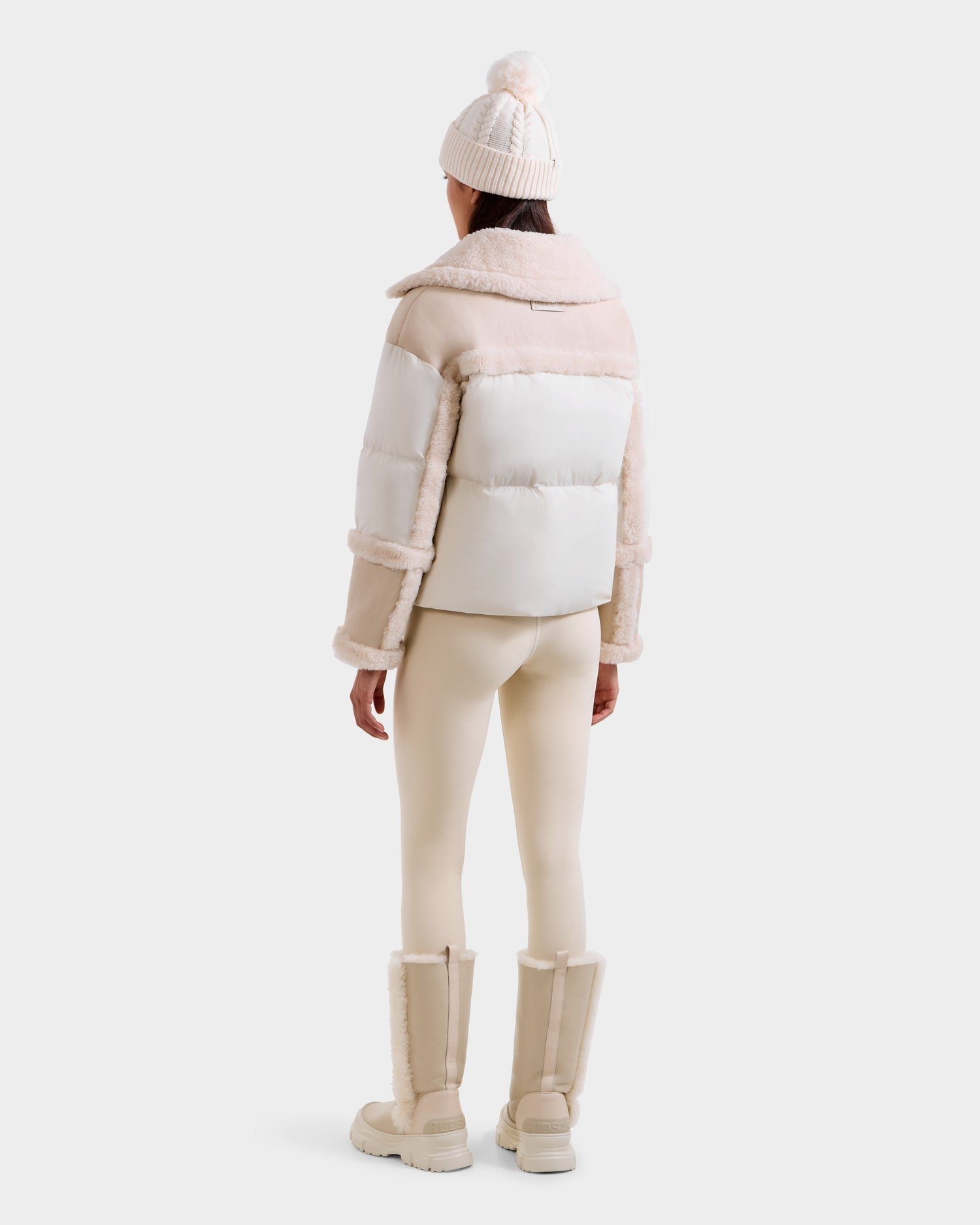 A person stands facing away in the Rudsak SHIRLEY - 8125613 Vanilla women's down jacket, paired with beige leggings, fur-lined boots, and a pom-pom knit beanie. A white background highlights this cozy winter look.