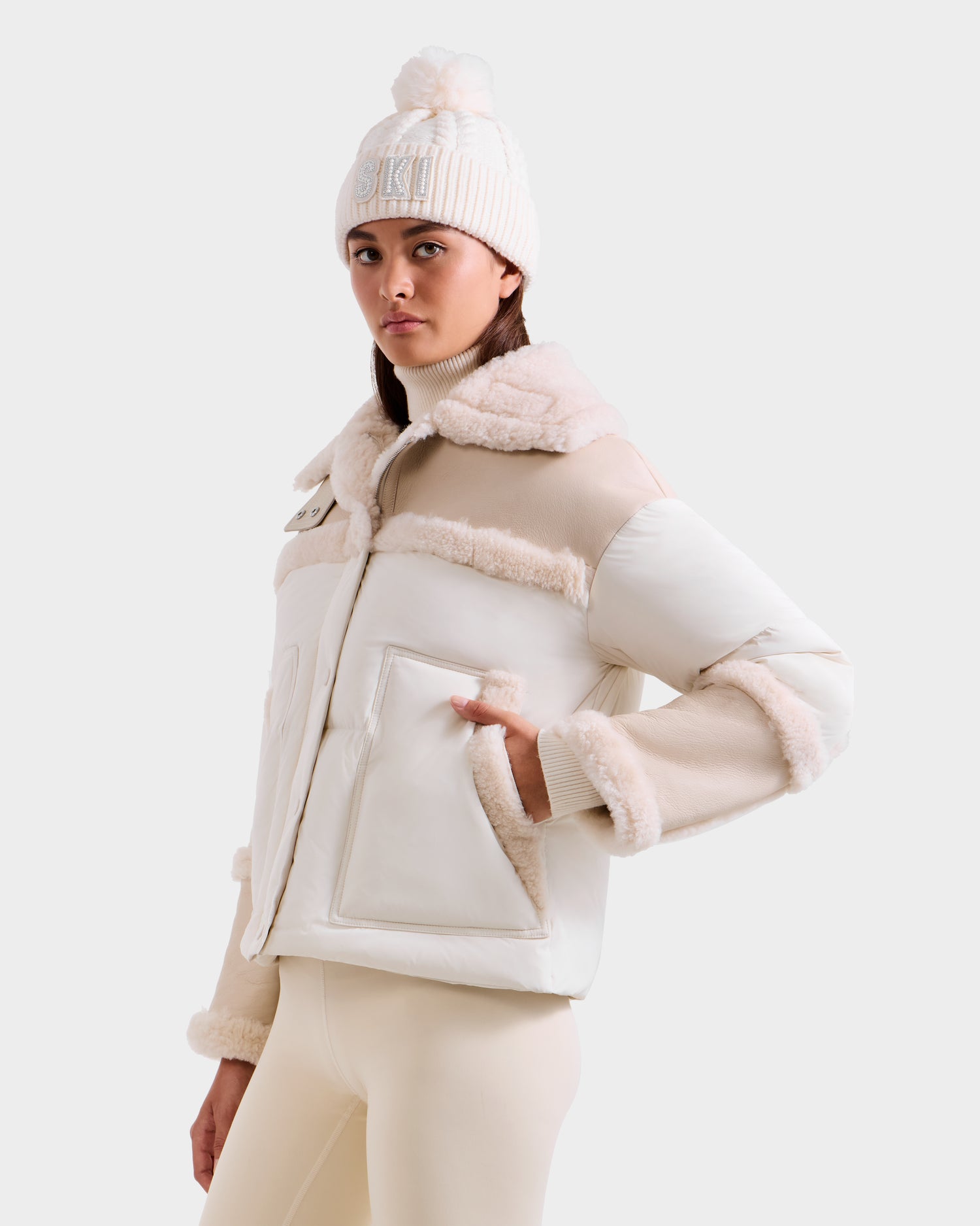 A woman poses facing forward with one hand in her pocket, wearing the Rudsak SHIRLEY - 8125613 Vanilla shearling jacket, a cream knit pom-pom hat, and light beige leggings against a plain white background highlighting her chic winter style.