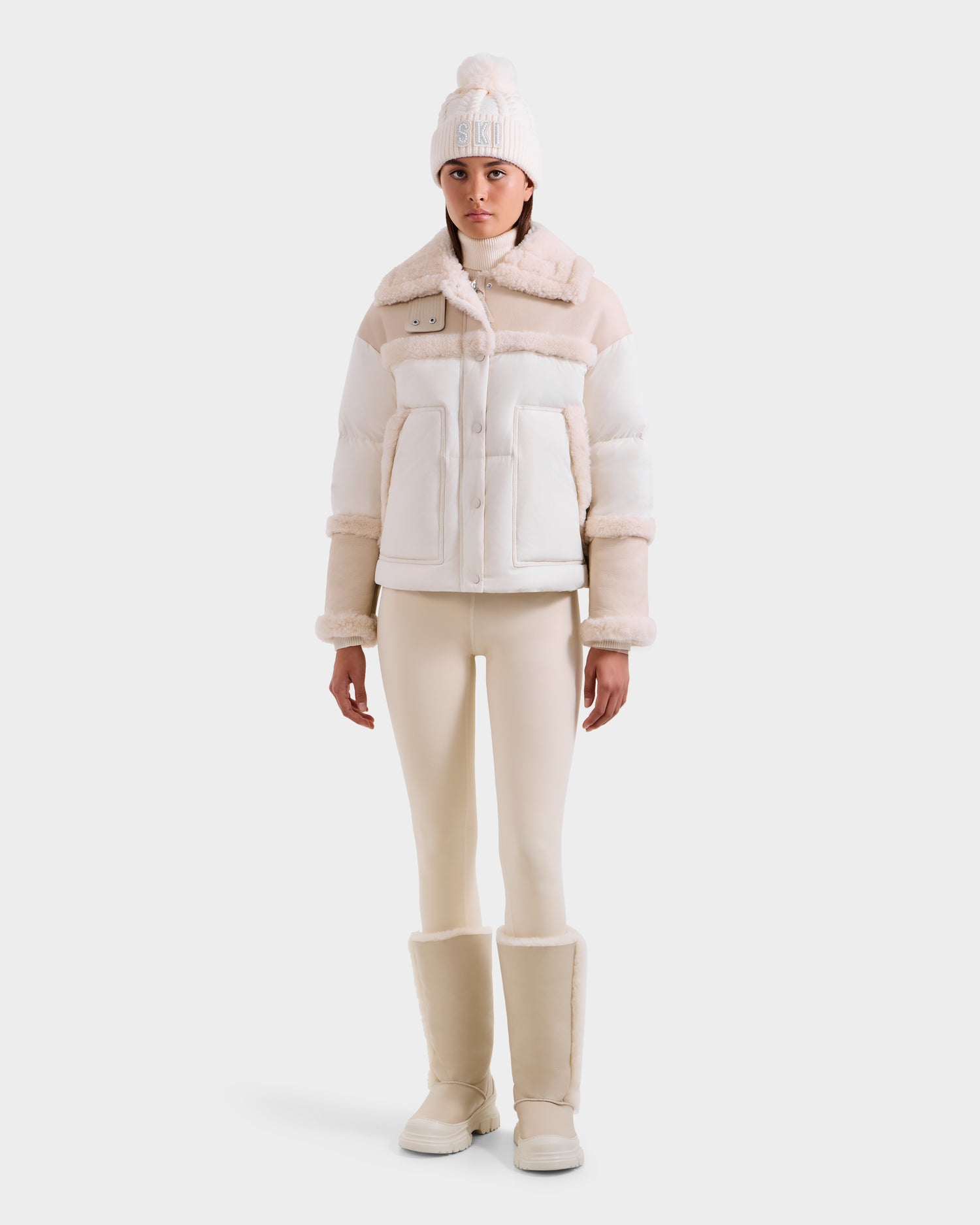 Dressed in the Rudsak SHIRLEY - 8125613 Vanilla shearling jacket, cream leggings, matching boots, and a white pom-pom knit hat, this effortlessly chic look is perfect for winter against a light background.