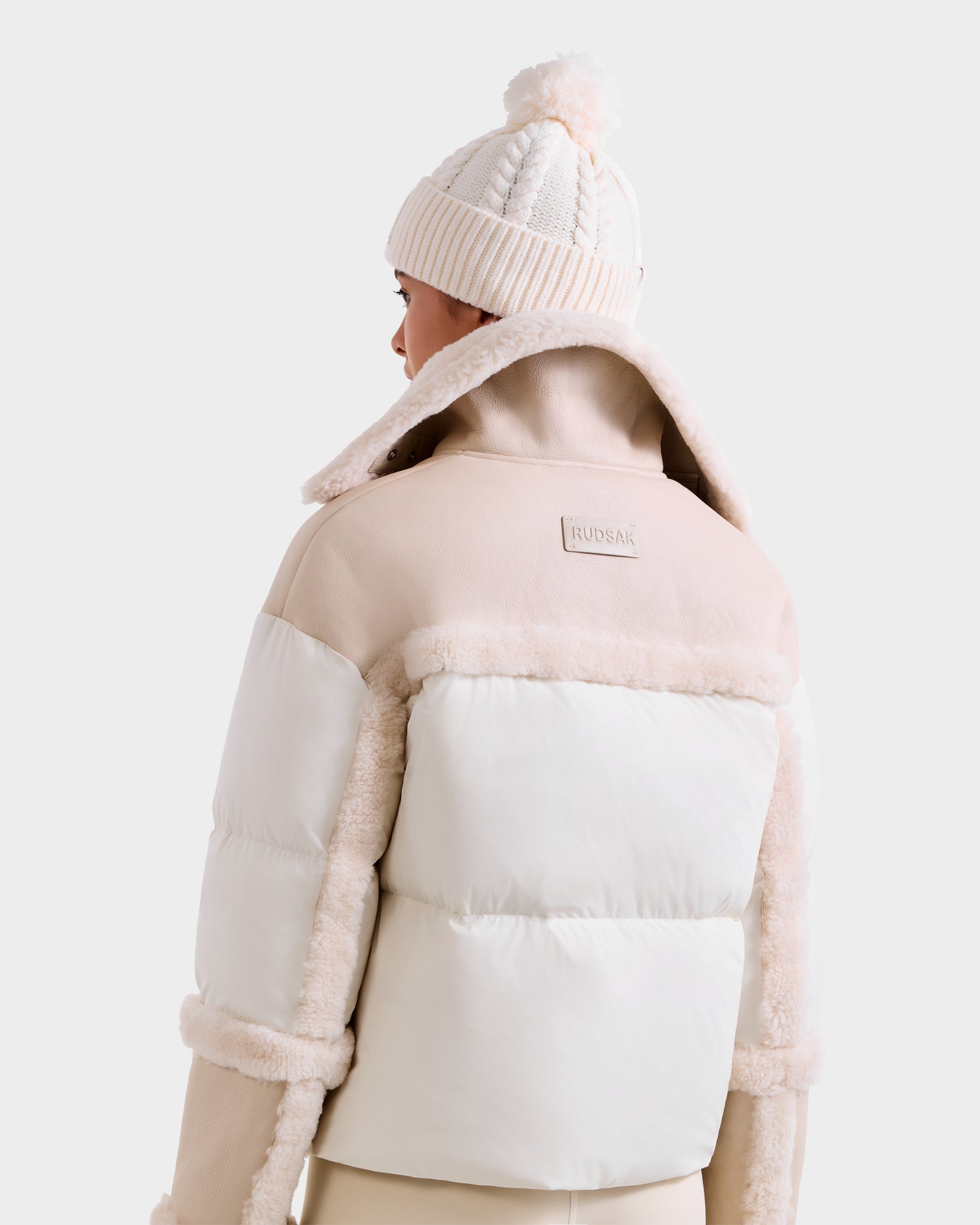 A person is shown from the back wearing the Rudsak SHIRLEY - 8125613 Vanilla women’s down jacket with faux fur trim and a cream knit beanie. The winterwear features a visible Rudsak label on the upper back.