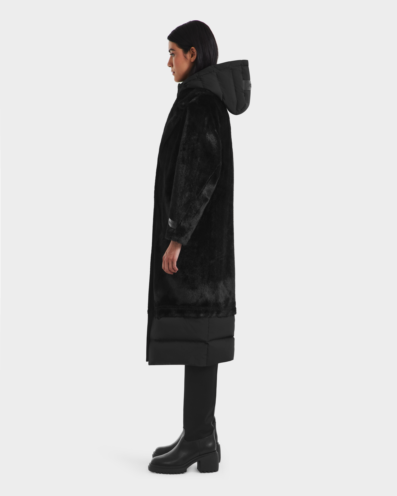 A person in profile wears the Rudsak SIENA - 8125542 Black down-insulated hooded coat, styled with black pants and ankle boots, set against a plain white background.