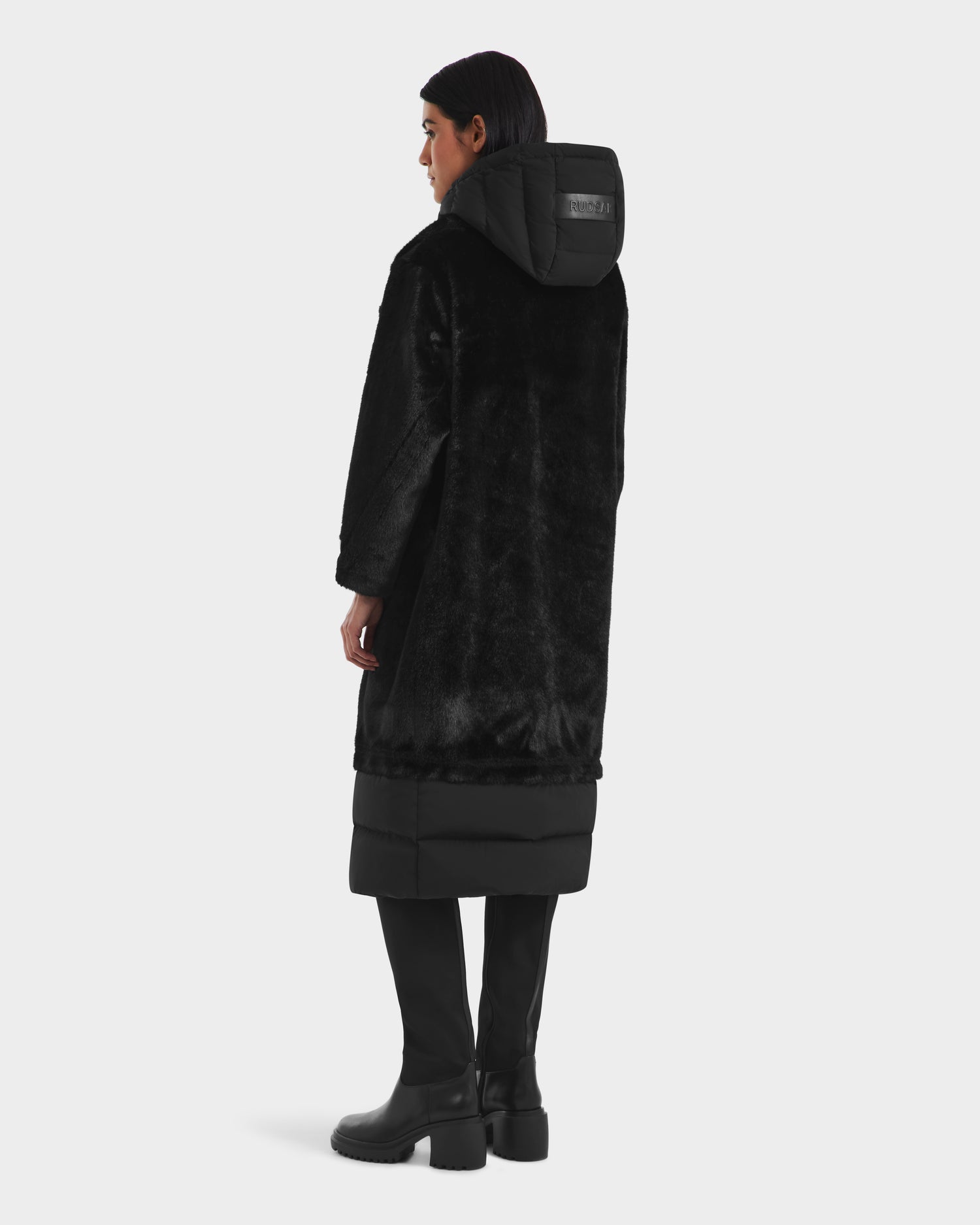 A woman faces away in a Rudsak SIENA - 8125542 Black faux fur winter coat with hood, leather patch detail, and quilted down-insulated hem, paired with black pants and boots against a plain white background.