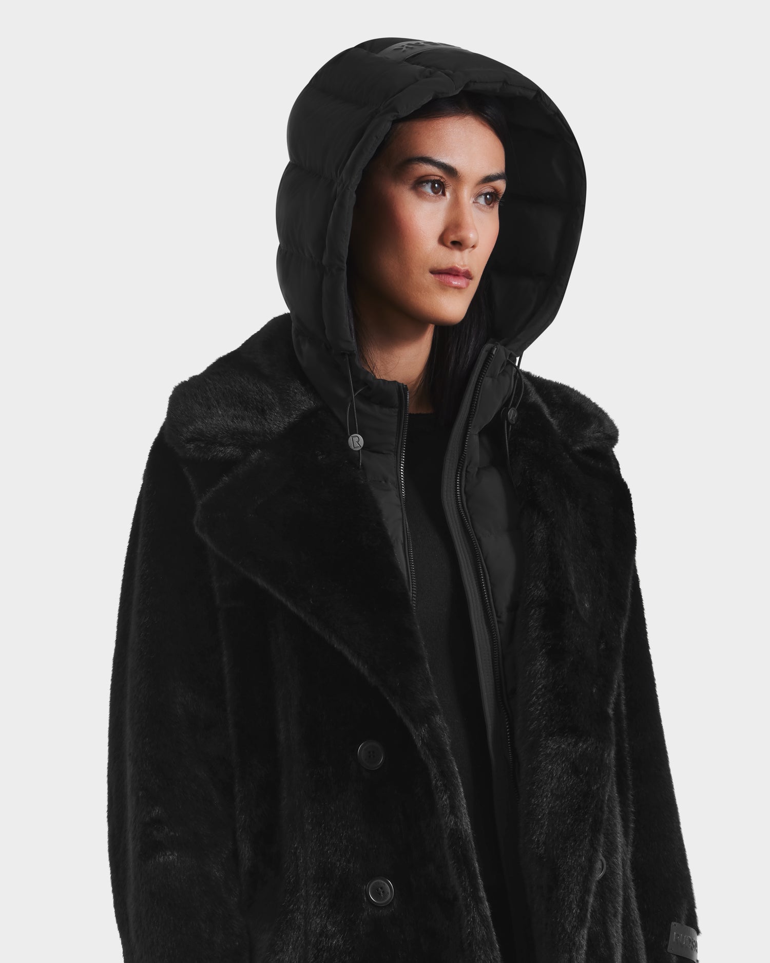 A woman wearing the Rudsak SIENA - 8125542 Black faux fur coat over a down-insulated layer looks ahead with a neutral expression against a plain white background.