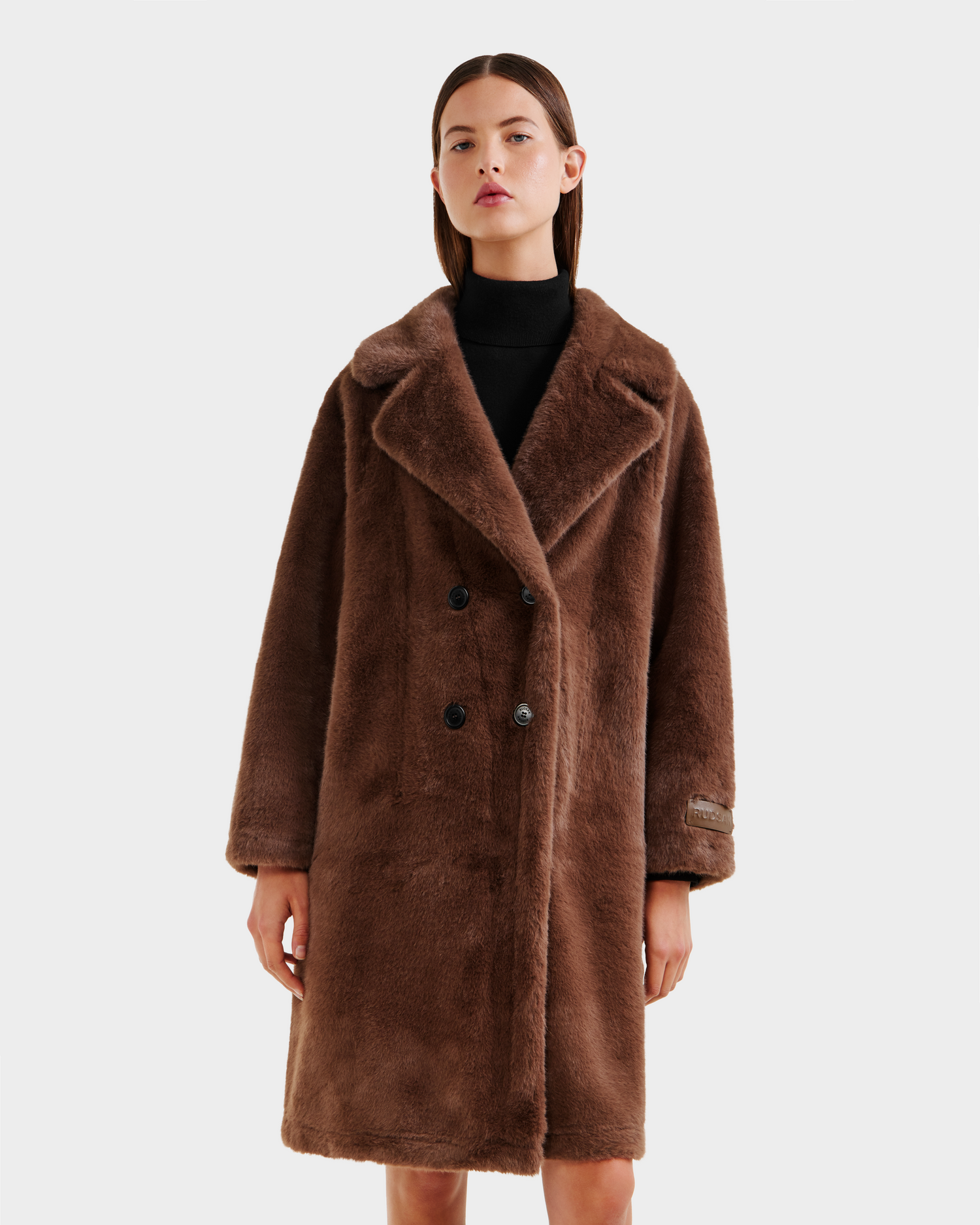 A woman stands facing forward against a white background, wearing the Rudsak SIENA - 8125542 Mocha, a long women’s faux fur coat with a wide collar and double-breasted buttons over a black turtleneck.