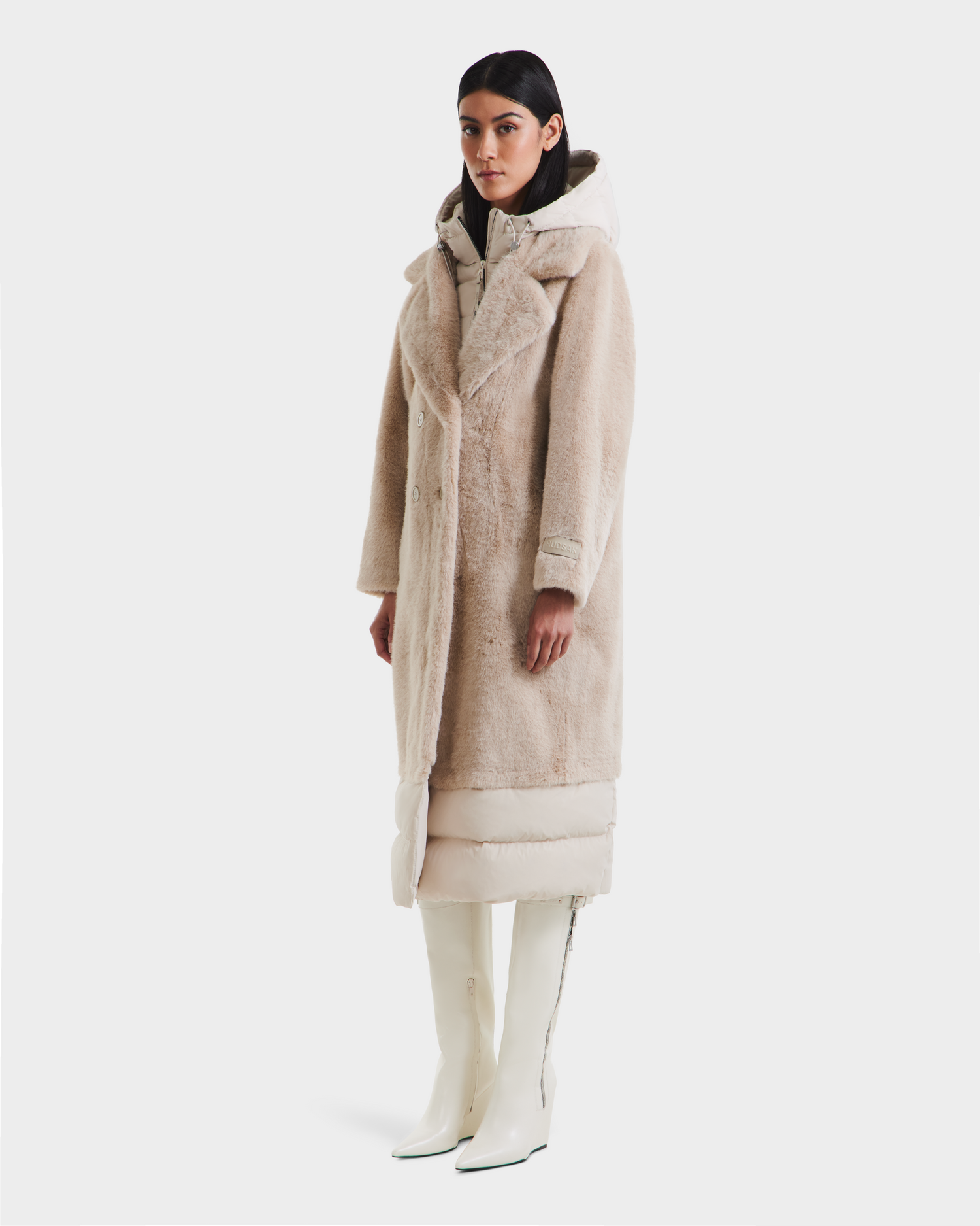 A woman faces forward against a white background, wearing the Rudsak SIENA - 8125542 Vanilla faux fur coat layered over a light puffer jacket and paired with white knee-high boots.
