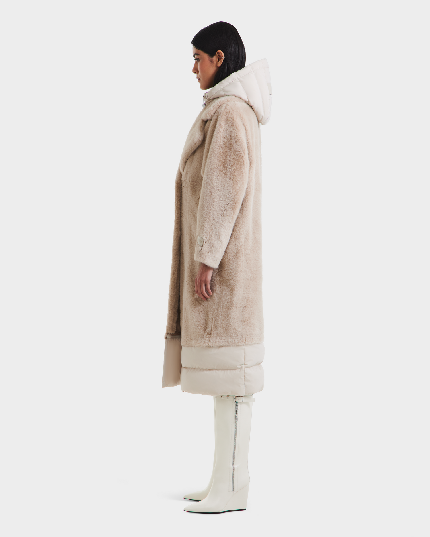 A woman stands in profile against a plain white background, wearing the Rudsak SIENA - 8125542 Vanilla women's faux fur coat with a hood, light-colored quilted hem detailing, and knee-high white boots with side zippers.