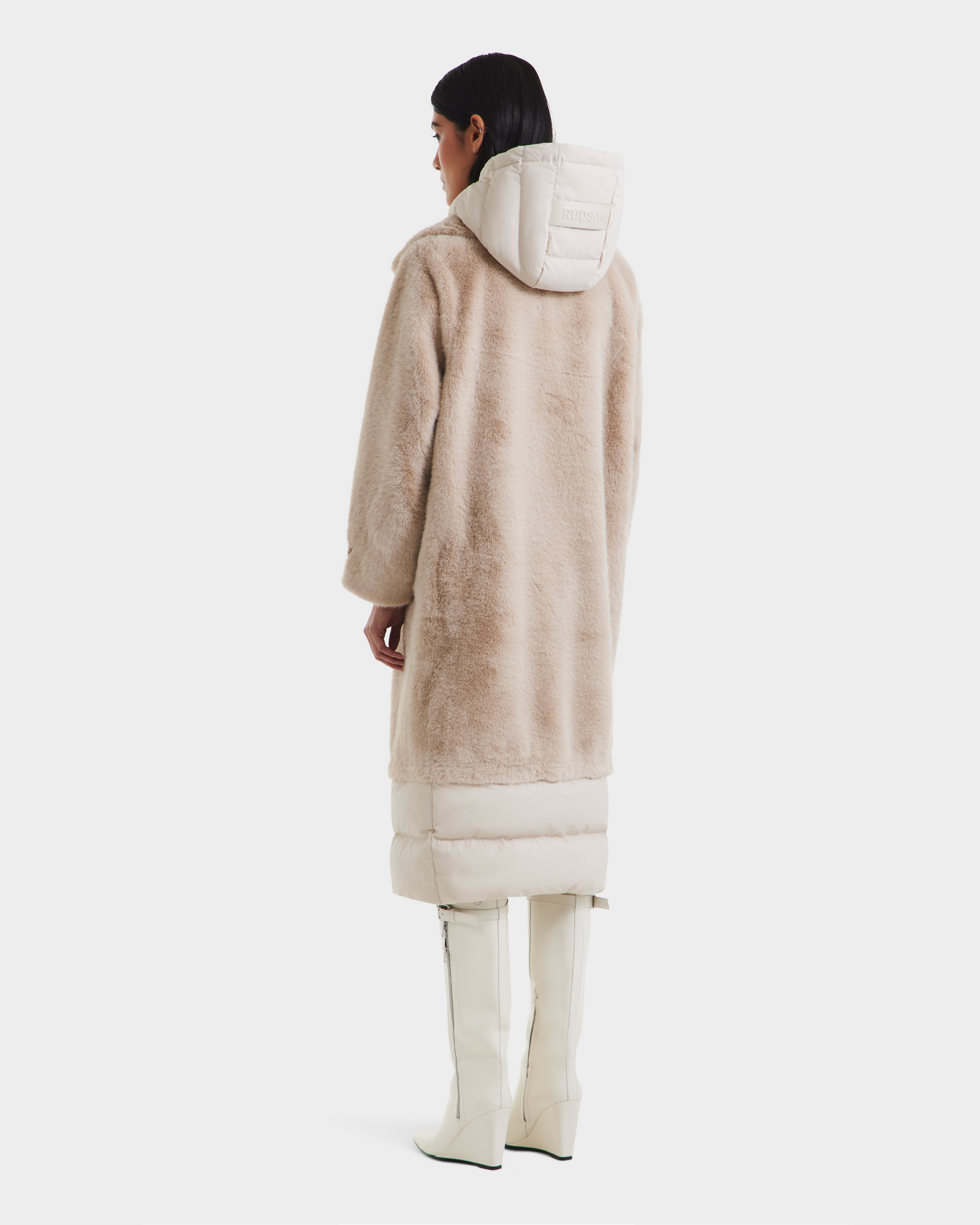A woman with straight dark hair wears the Rudsak SIENA - 8125542 Vanilla, a long beige faux fur coat with padded hood and lower section, paired with white zippered knee-high boots against a plain white background.