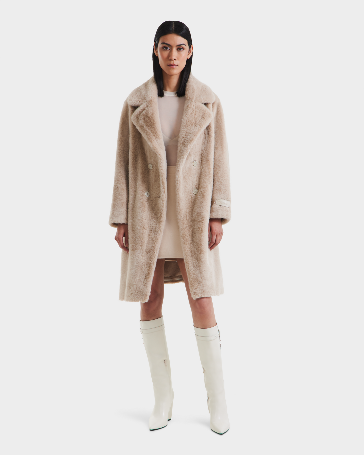A woman faces forward in the Rudsak SIENA - 8125542 Vanilla faux fur coat over a pale dress and knee-high white boots, set against a plain white background.