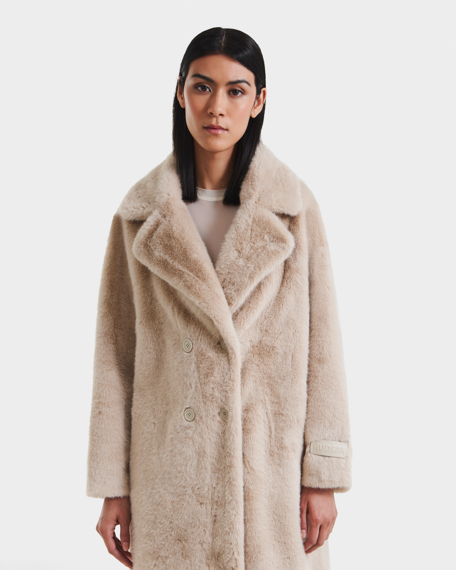 A woman with straight black hair models the Rudsak SIENA - 8125542 Vanilla, a beige double-breasted women’s faux fur coat, over a sheer white top against a plain white background.