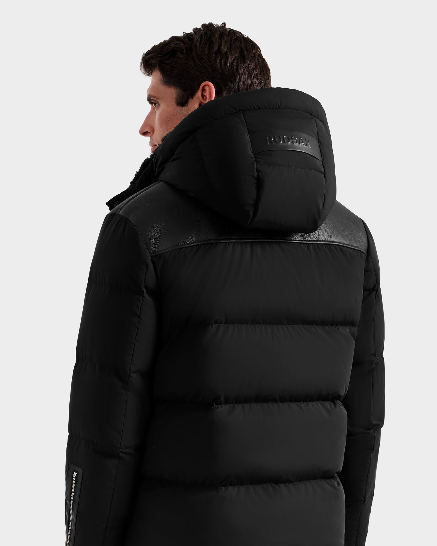 A man is seen from behind wearing the Rudsak SIMON - 6125597 Black men’s down puffer jacket, featuring leather-like shoulder panels and an embossed RUDSAK hood, standing out against a plain, light background.